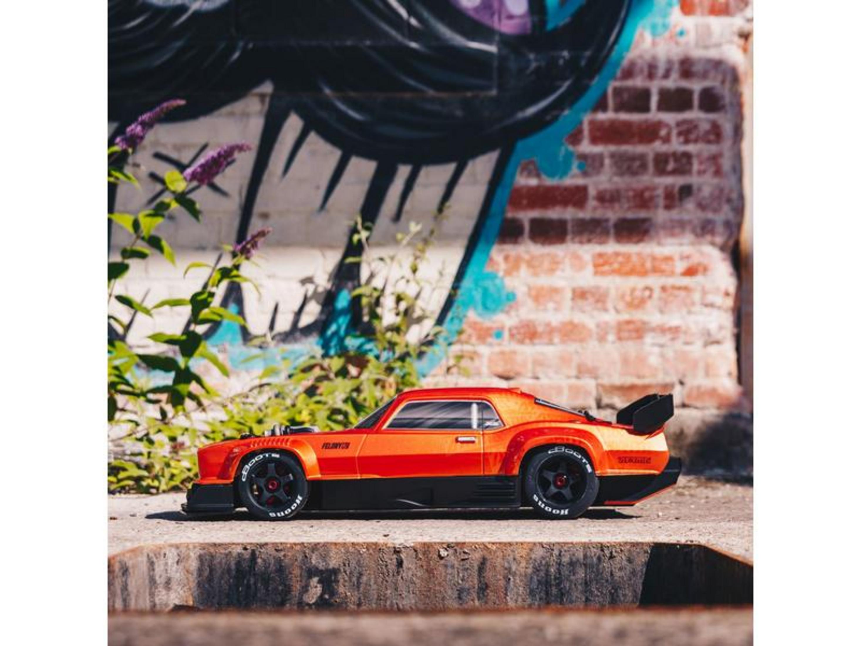 Un coche RC naranja con alerón negro sobre bloque de hormigón, con muro de grafitis detrás.