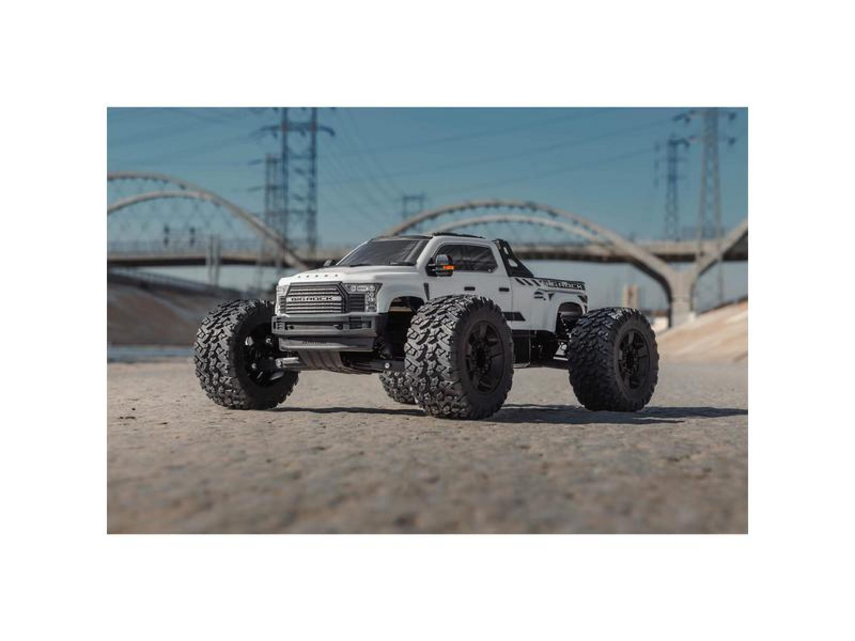 Un monster truck blanco con llantas grandes está sobre suelo arenoso, puentes al fondo.