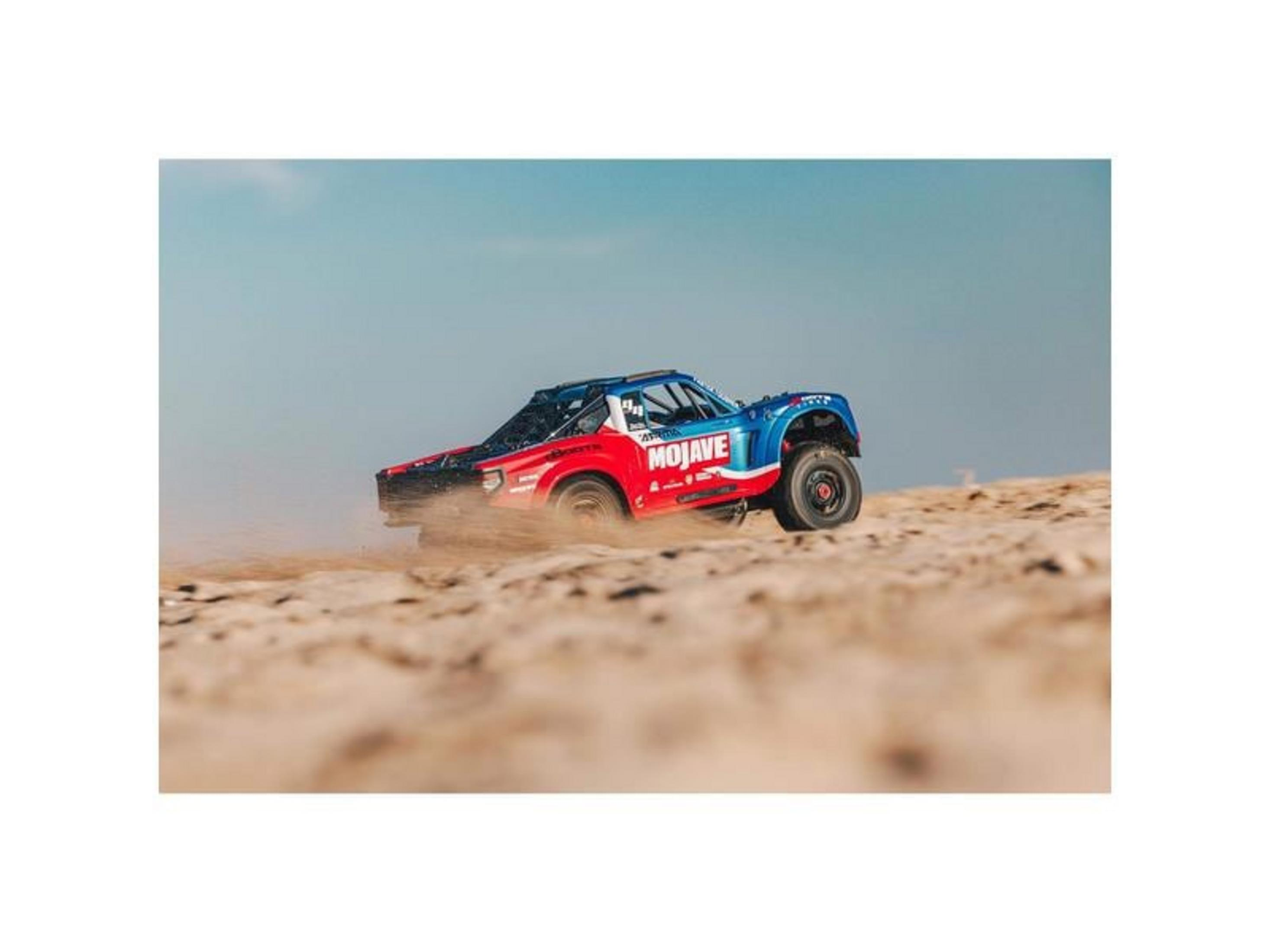 Ein blau-rotes RC-Offroad-Auto fährt auf sandigem Gelände und wirbelt Staub auf.