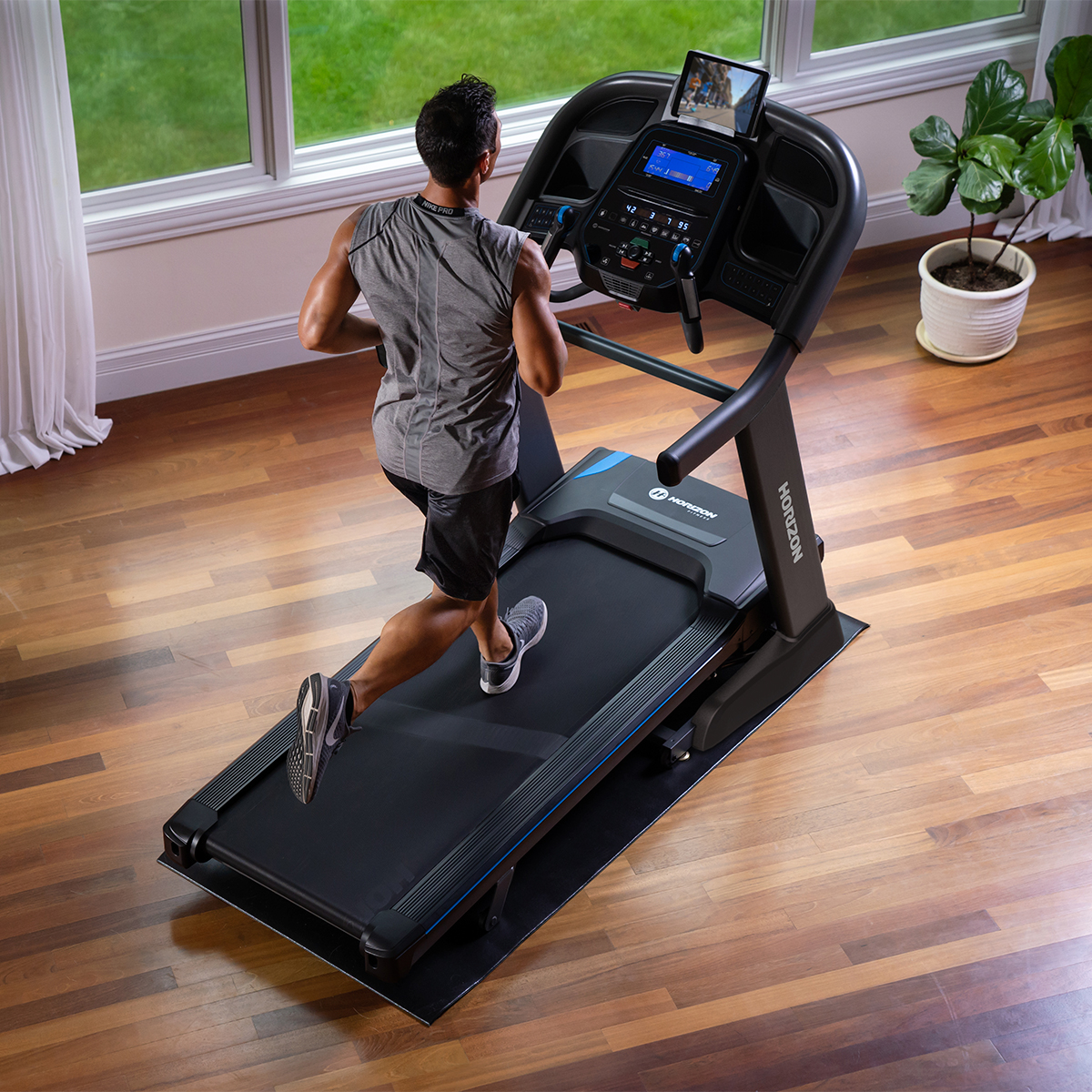 Hombre corriendo en cinta de correr en sala con suelo de madera y ventanas.