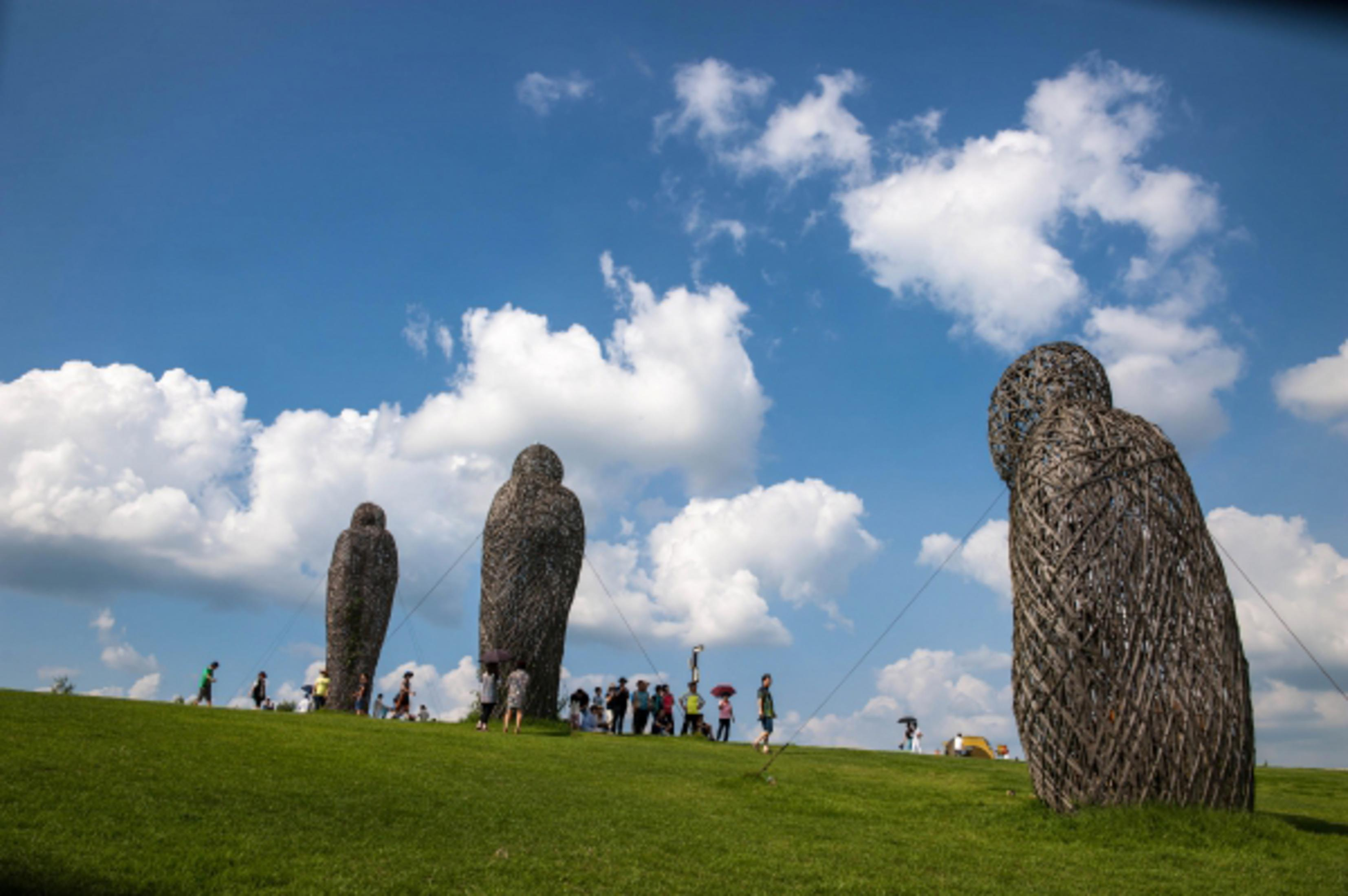 Tre grandi sculture simili a rami si ergono su una collina erbosa sotto un cielo blu.