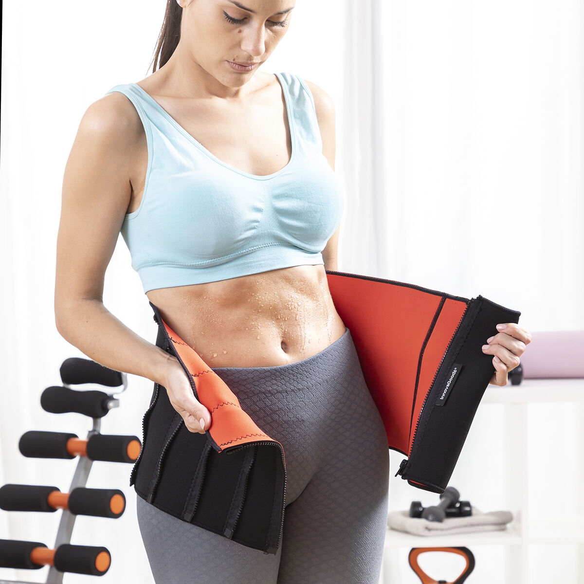 Frau mit Schweiß auf dem Bauch hält einen orangefarbenen Neopren-Taillengürtel.