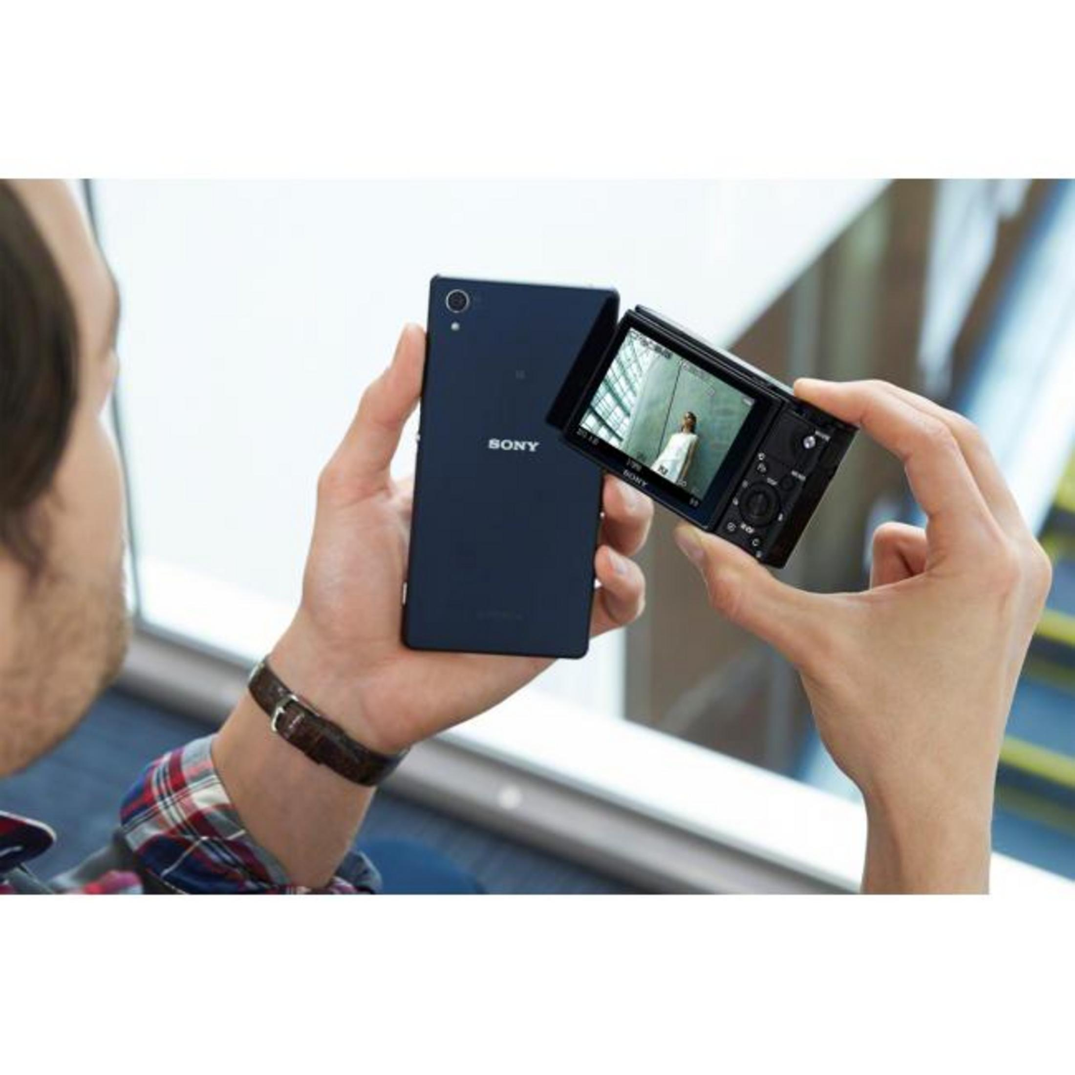 Mann hält Smartphone und Kamera. Person trägt kariertes Hemd und Armband.