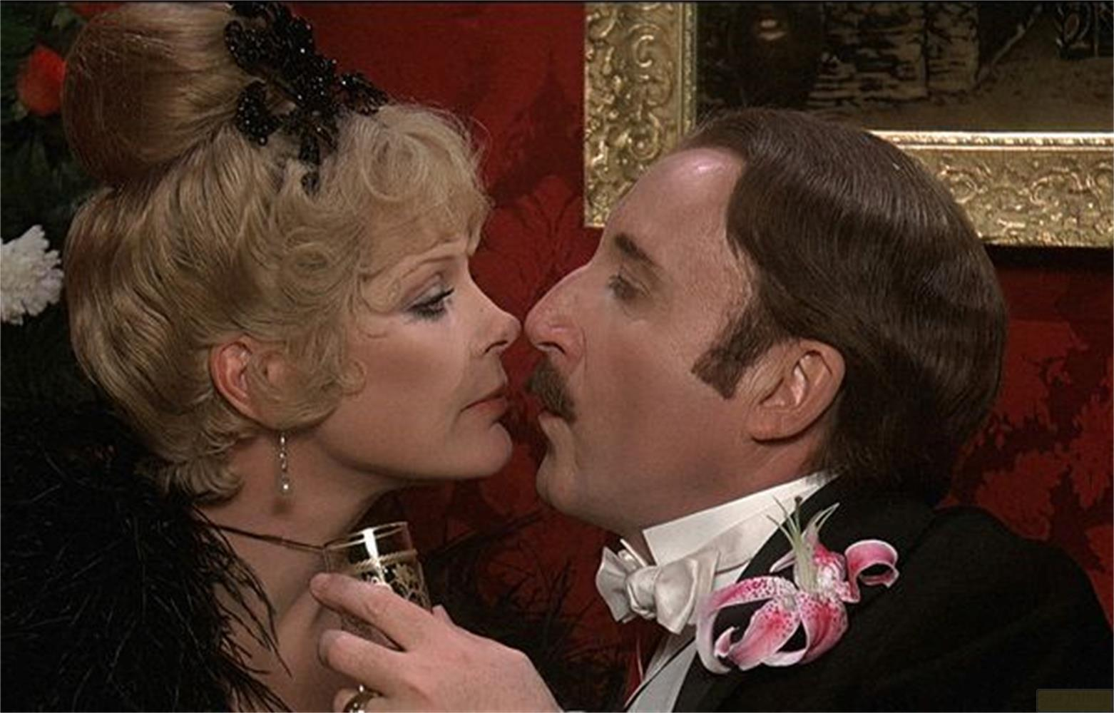 Uomo e donna si baciano, donna tiene bicchiere, uomo indossa fiore sull'abito.