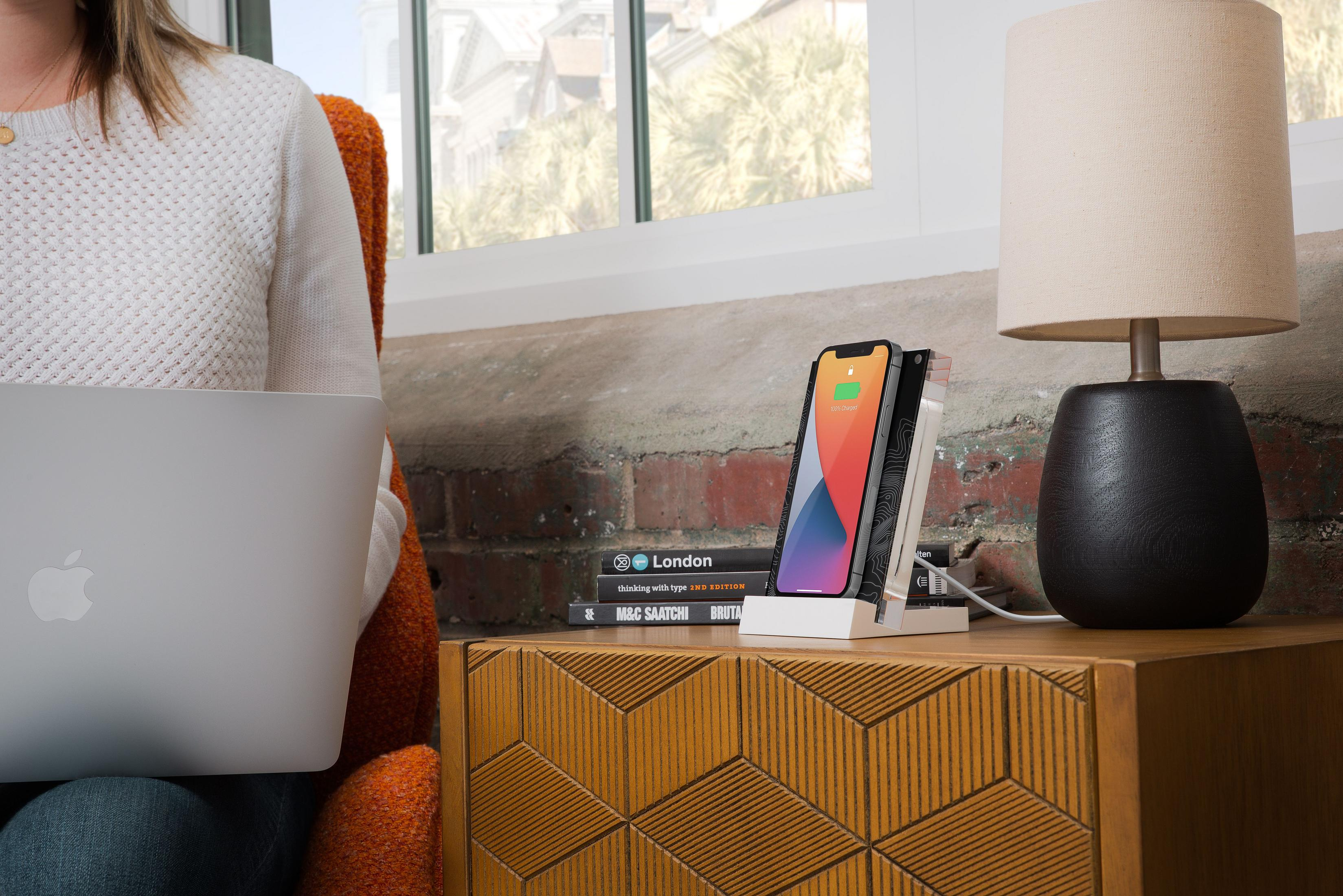Personne avec ordinateur portable, téléphone sur chargeur, livres et lampe sur commode.