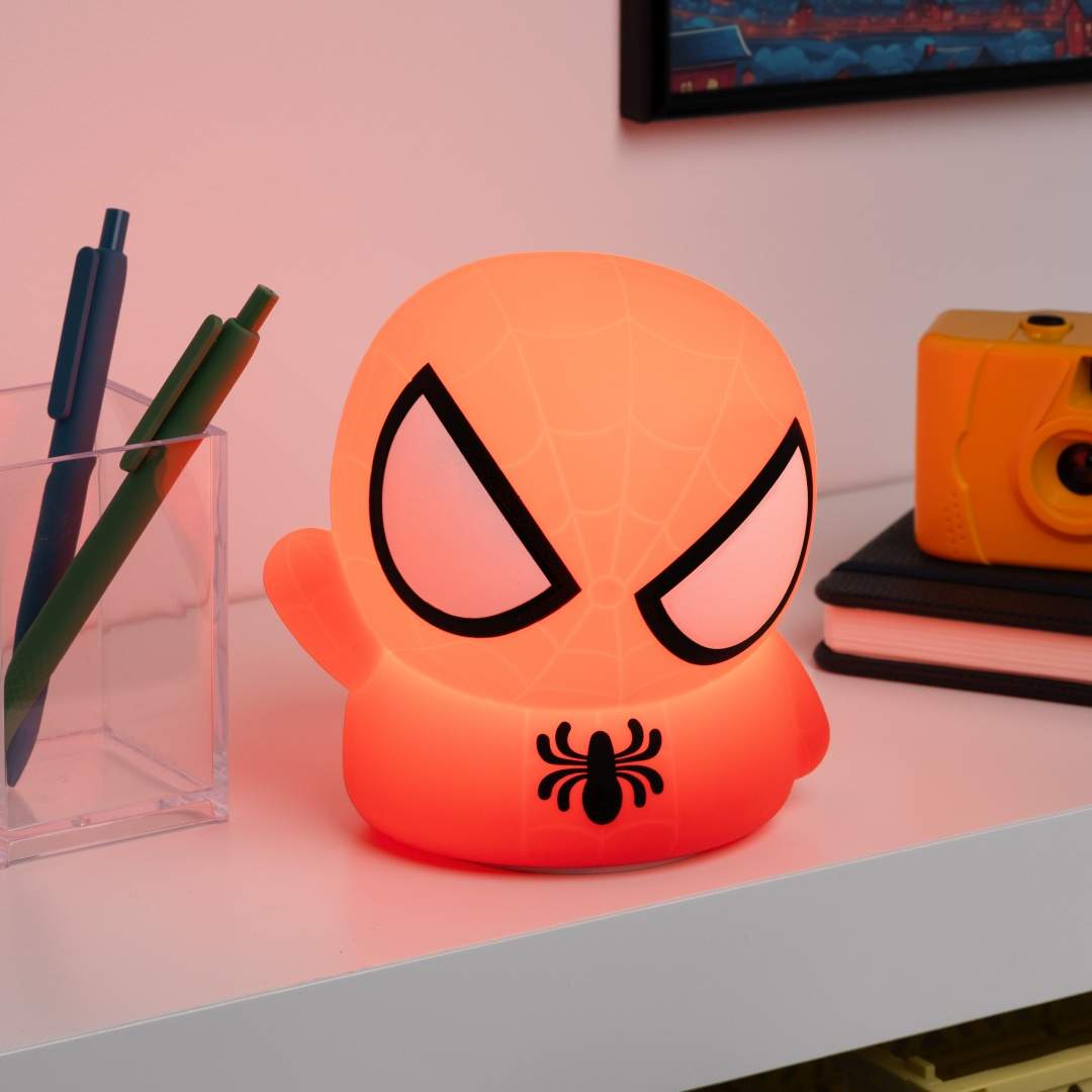 Świecąca pomarańczowa lampka w stylu Spidermana stoi na białej półce z długopisami i aparatem.