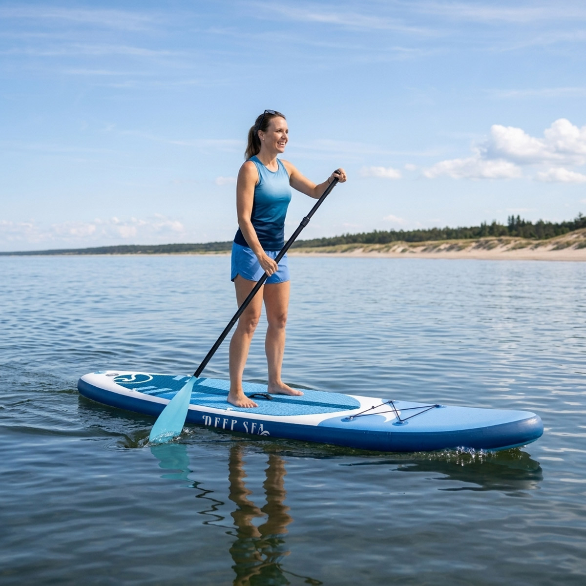 Kobieta pływa na desce SUP po spokojnej wodzie, plaża i drzewa w tle.