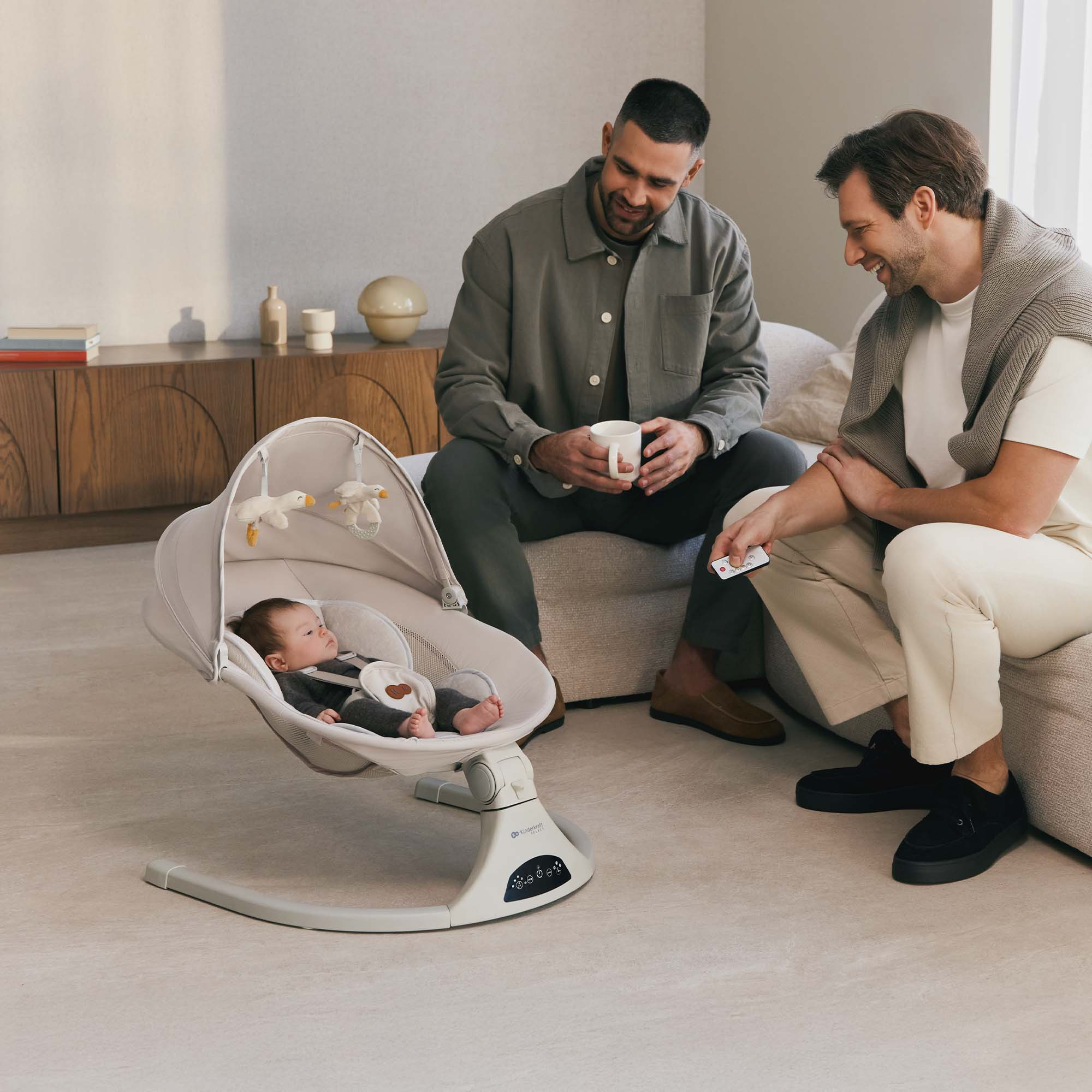 Zwei Männer sitzen neben einem Baby in einer Wippe. Einer hält eine Tasse, der andere eine Fernbedienung.