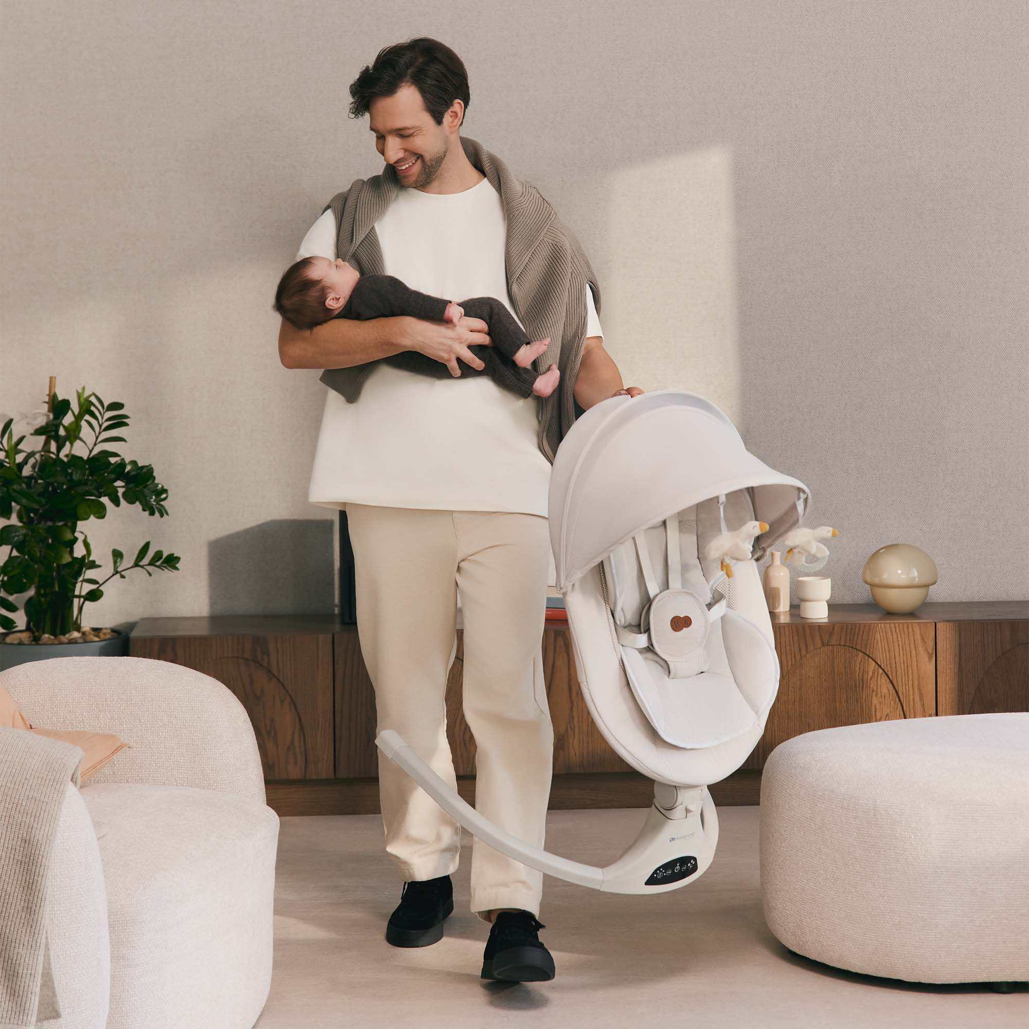 Lächelnder Mann hält schlafendes Baby, trägt weiße Babyschaukel. Wohnzimmer.