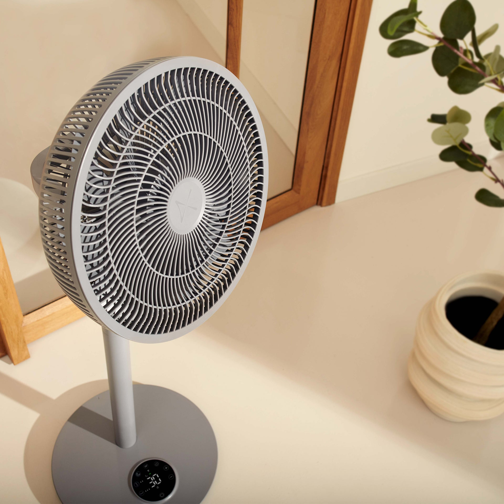 Ein grauer oszillierender Ventilator mit Digitalanzeige steht auf einem cremefarbenen Boden neben einer Topfpflanze.