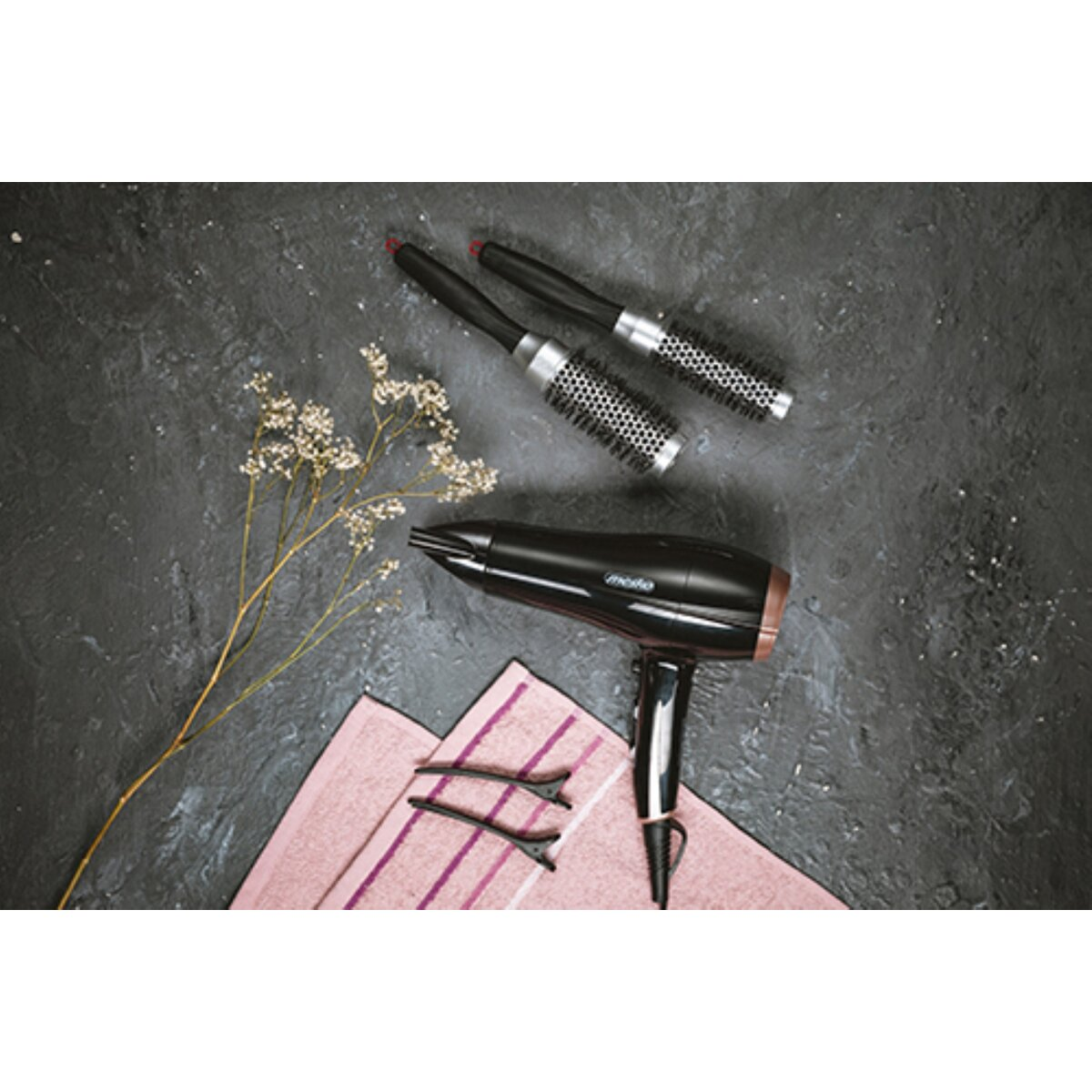 Ein schwarzer Haartrockner mit zwei Bürsten, Haarklammern und getrockneten Blumen auf einem rosa Handtuch.