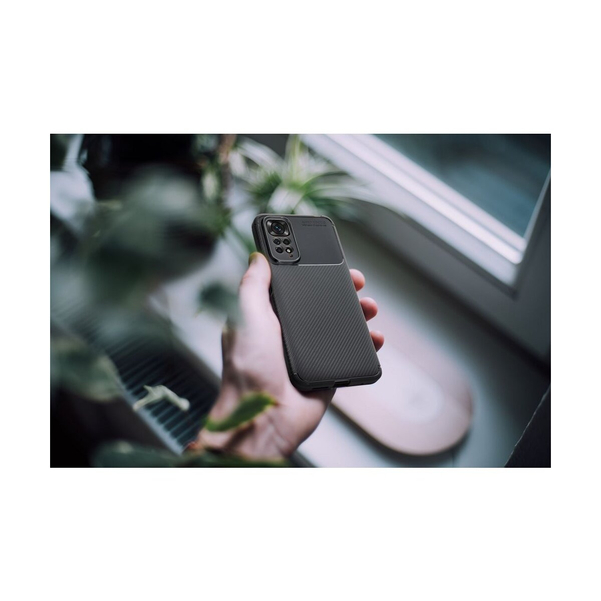 Eine Hand hält ein dunkles Handy mit Karbonmuster und Kameramodul. Pflanzen im Vordergrund.