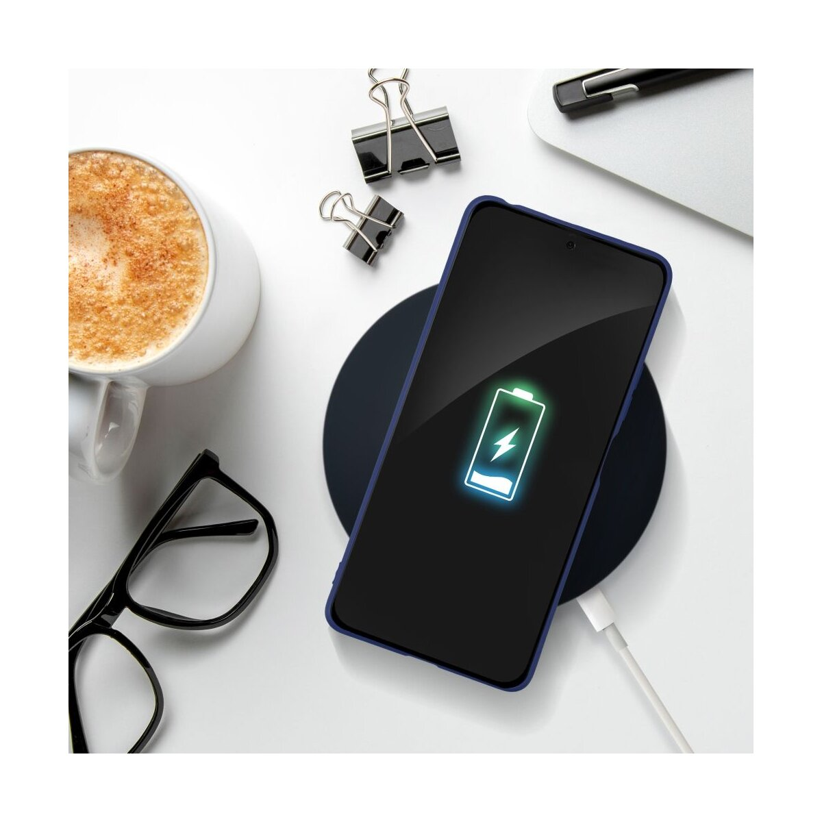 Ein Smartphone mit Ladeanzeige auf einem kabellosen Ladegerät, daneben Kaffee, Brille und Büroklammern.