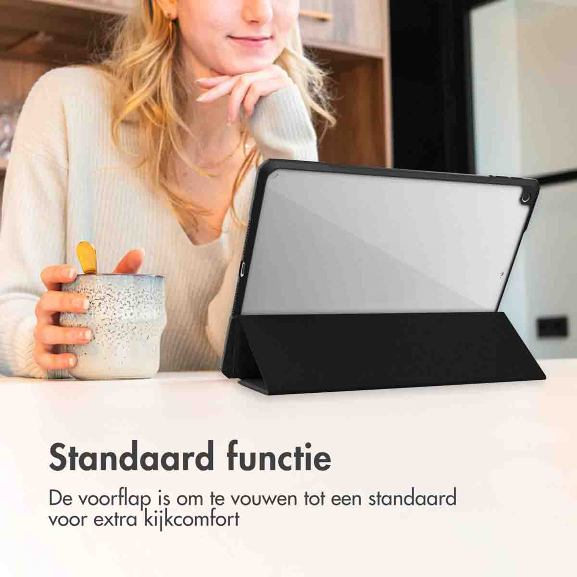 A woman holds a mug while a tablet is propped up on a stand.