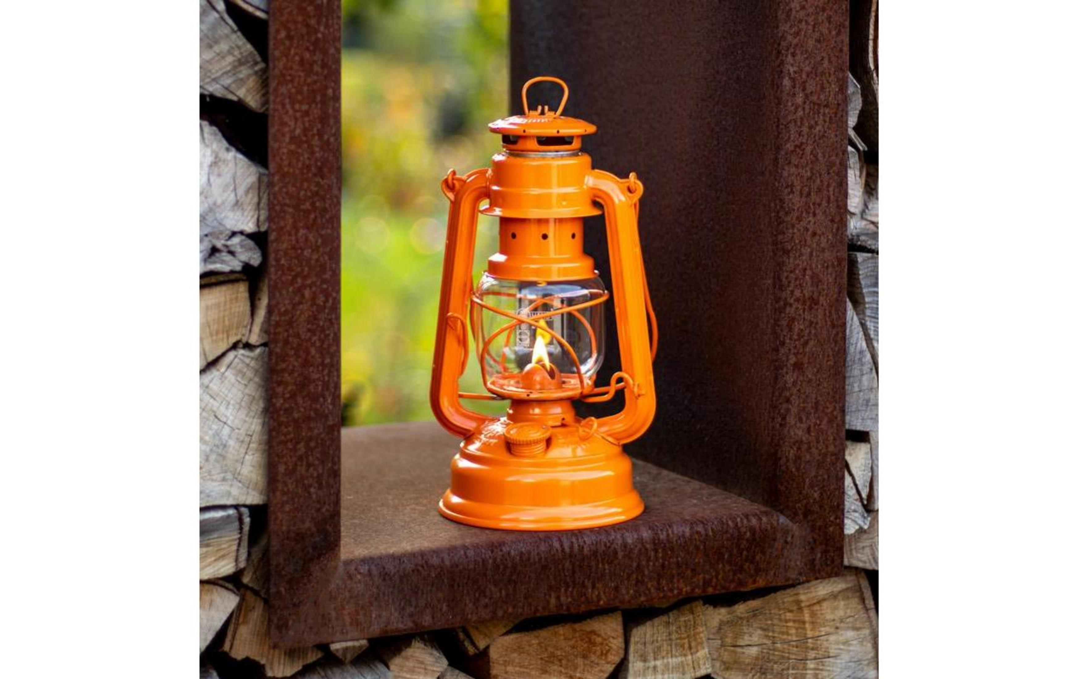 Eine orangefarbene Laterne brennt auf einem rostigen Regal, umgeben von Holzscheiten.