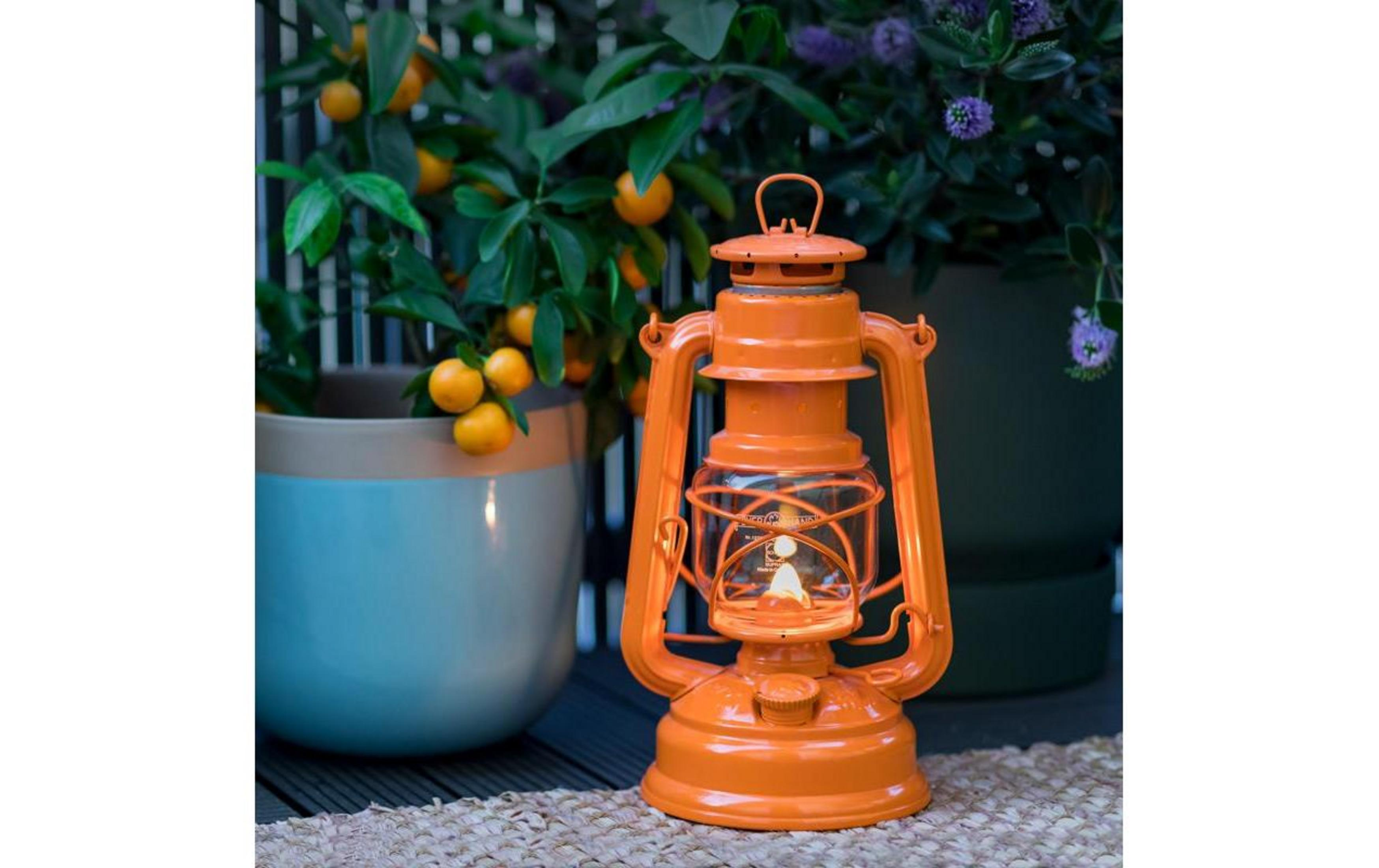 Eine orangefarbene Laterne steht vor einem Topf mit Orangen und lila Blumen.