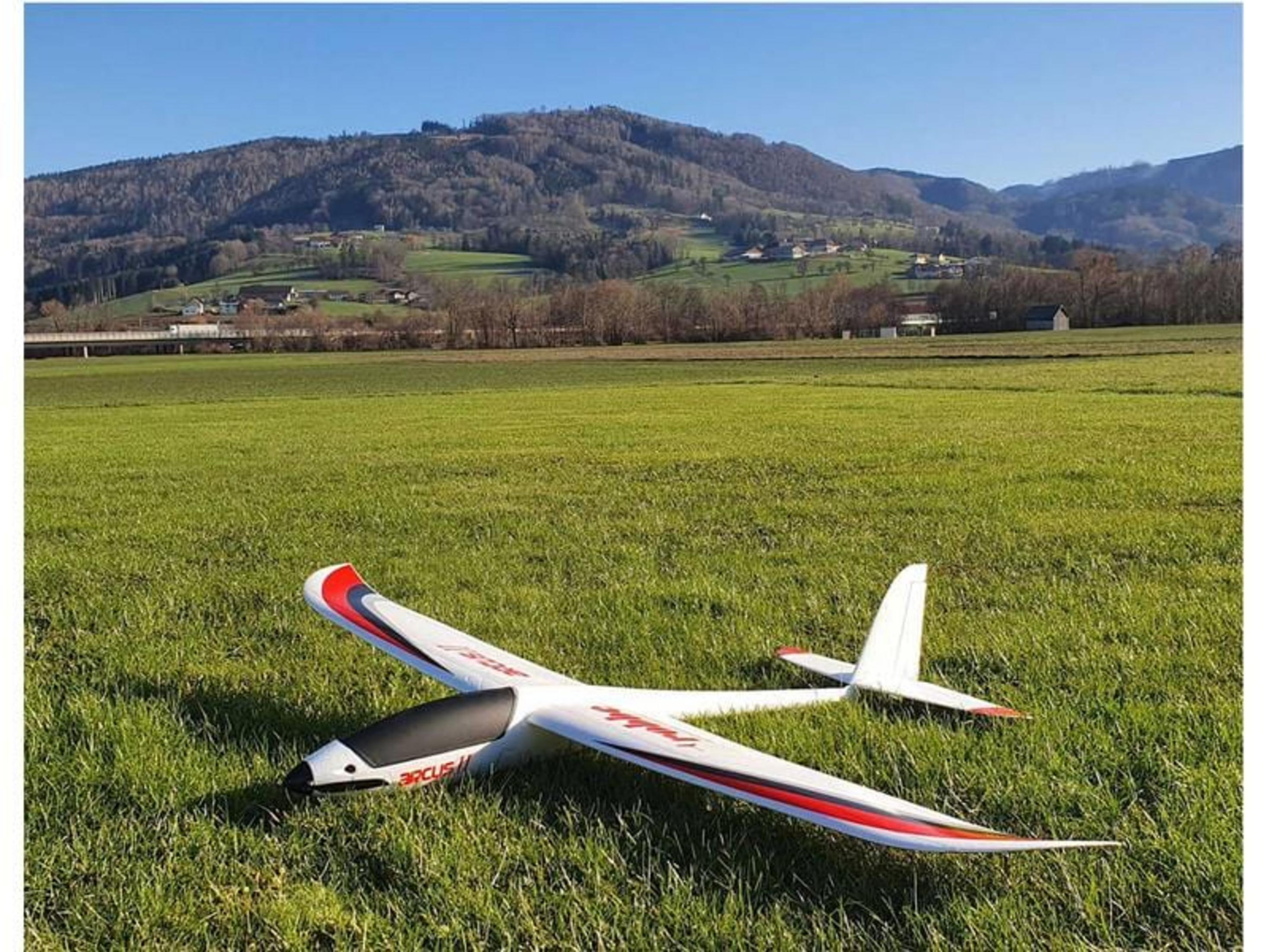 Un planeur blanc aux accents rouges repose sur l'herbe. Montagnes en arrière-plan.
