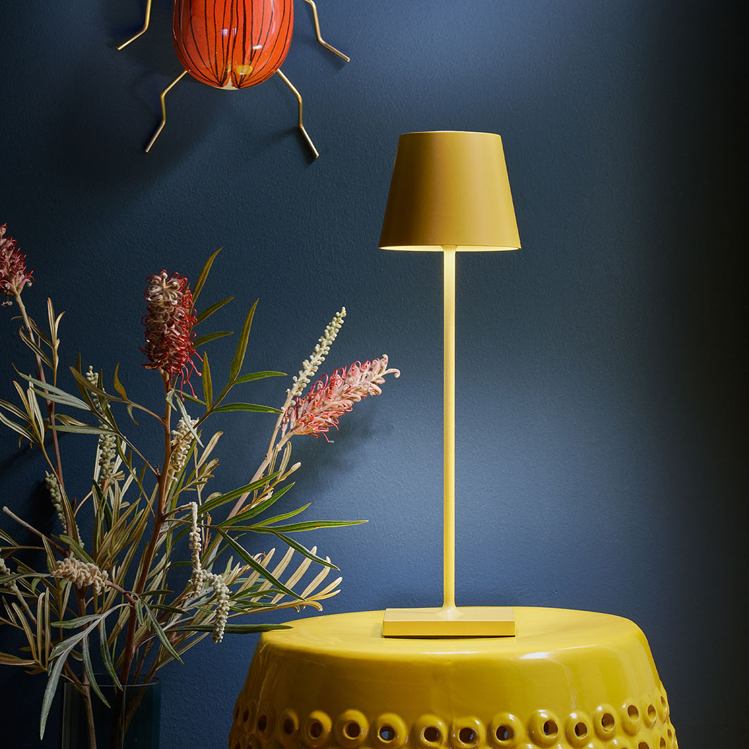 Eine gelbe Tischlampe steht auf einem gelben Hocker, daneben Blumen. An der Wand ein rotes Insekt.