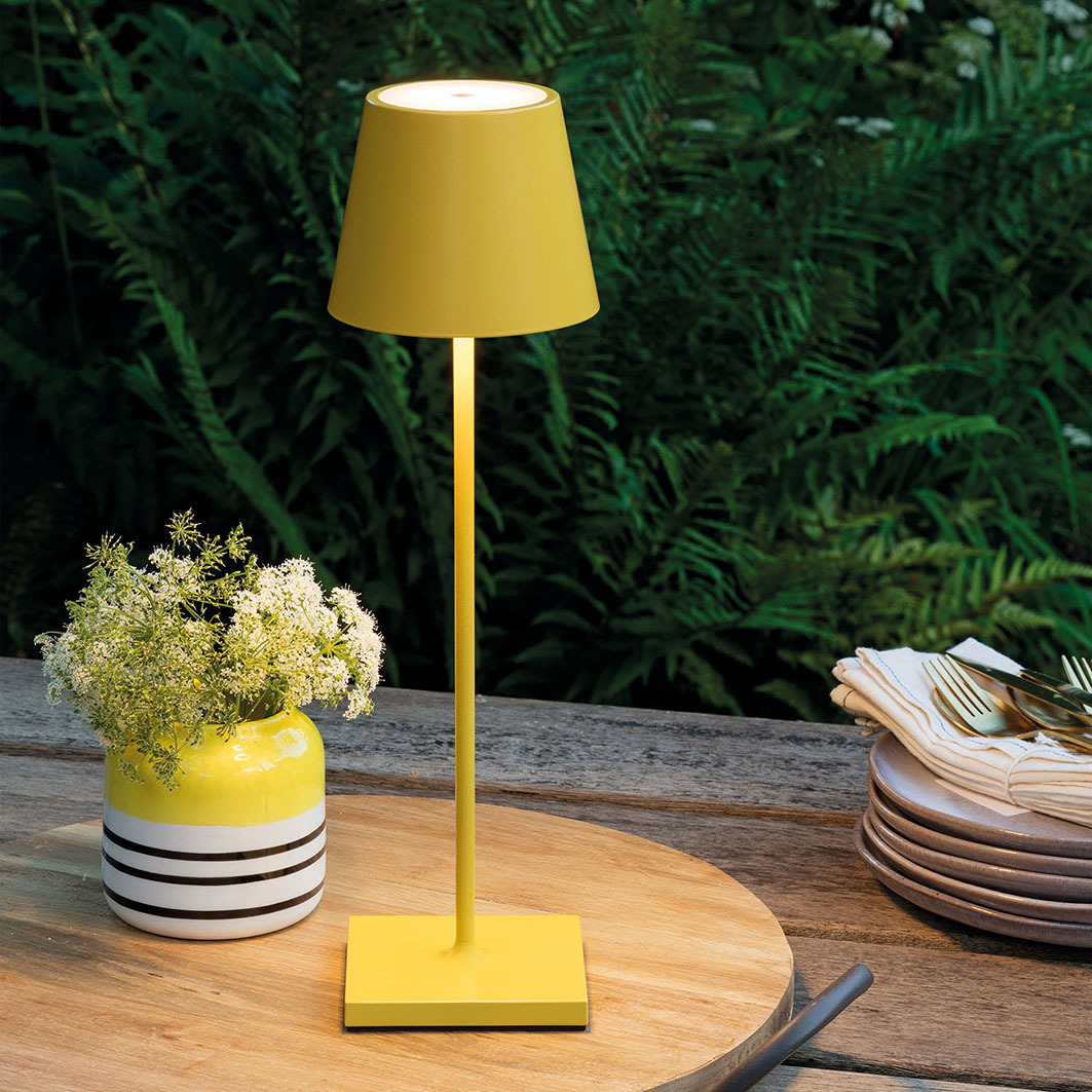 Eine gelbe Tischleuchte steht auf einem Holztisch neben einer Vase mit Blumen.