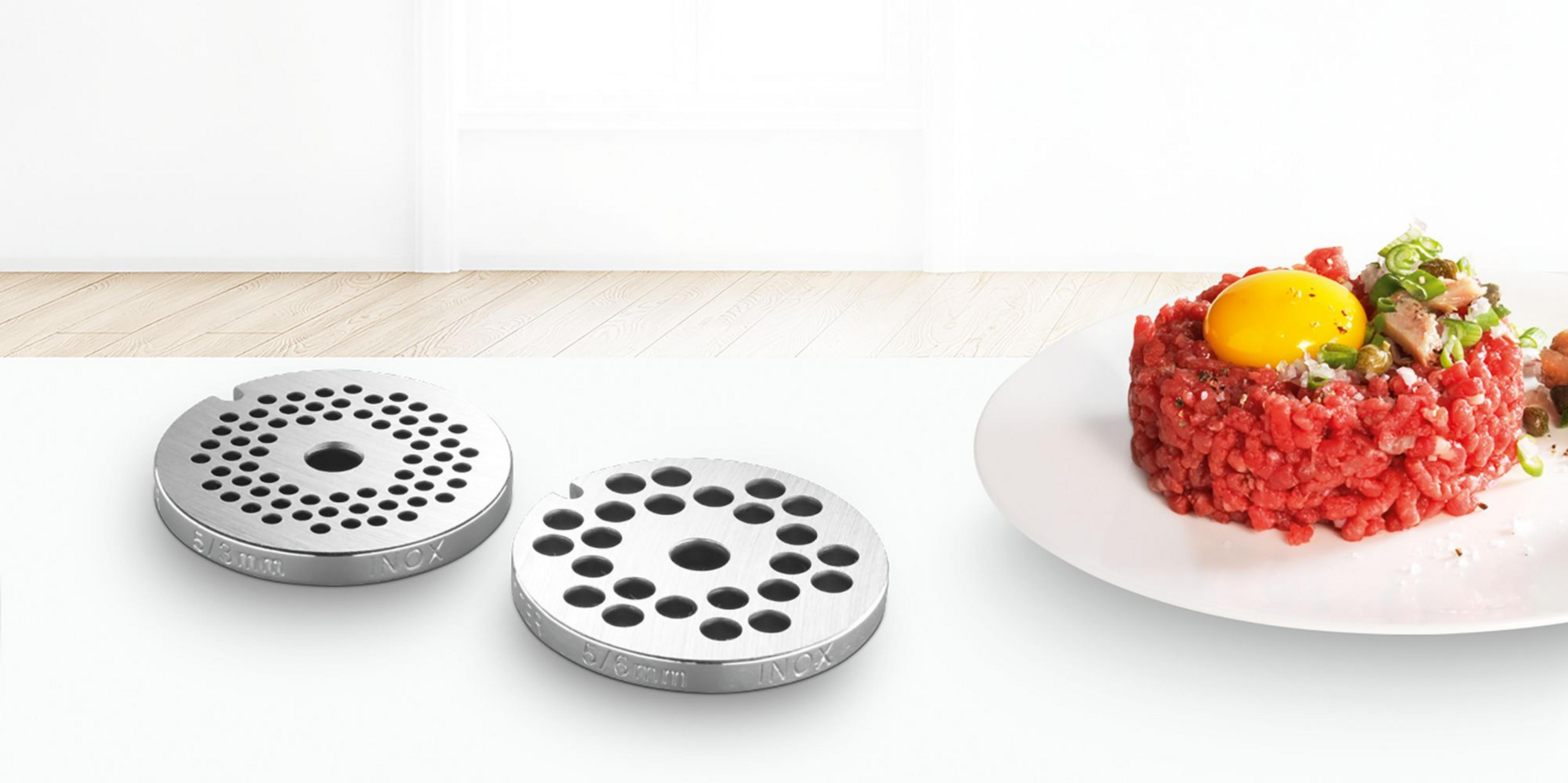 Zwei Metall-Fleischwolfscheiben liegen neben einem Teller mit rohem Rinderhackfleisch-Tartar.