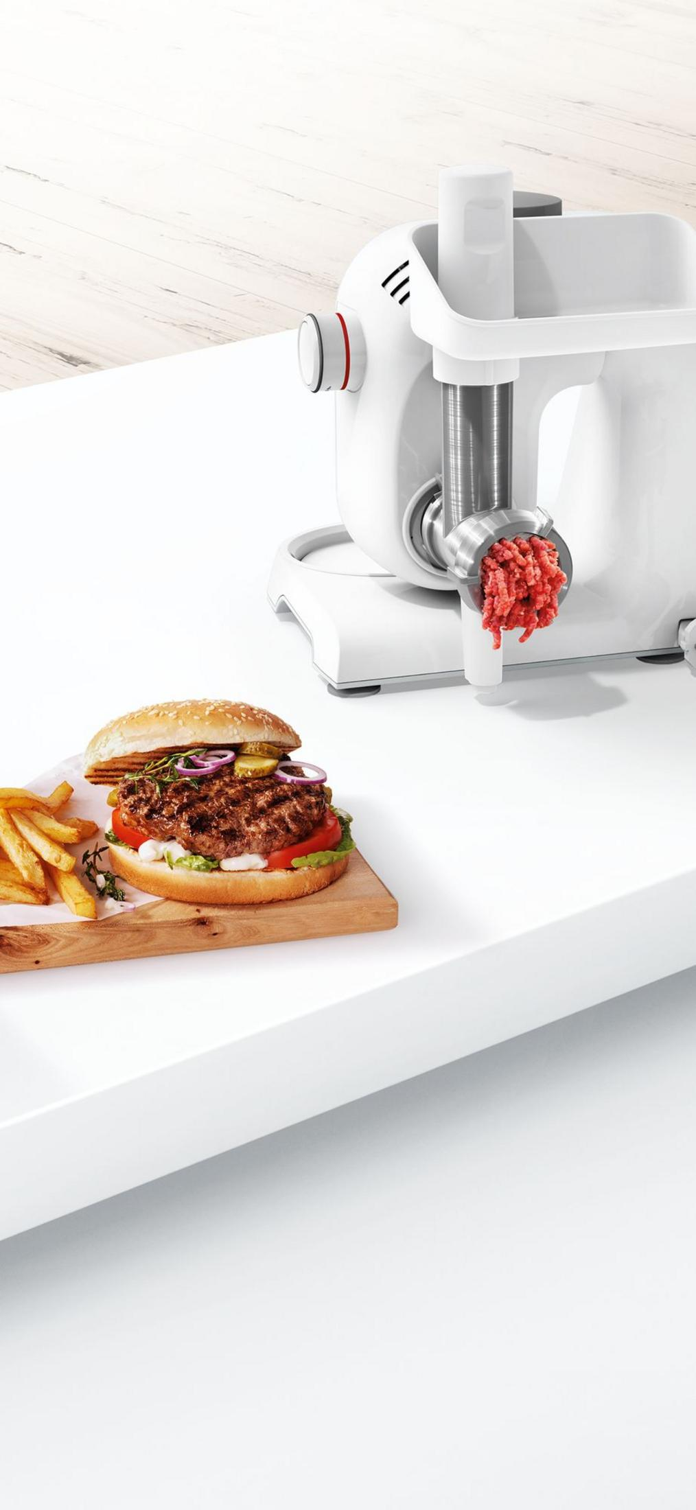 Fleischwolf mit Hackfleisch, daneben ein Burger mit Pommes auf einem Holzbrett.