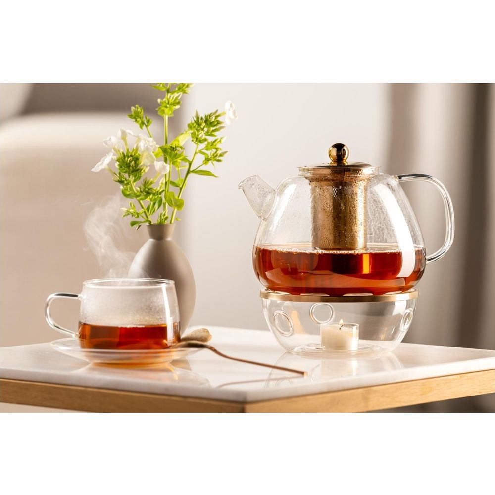 Eine Glasteekanne mit Tee steht auf einem Wärmer mit Kerze. Eine Teetasse und Blumen sind in der Nähe.
