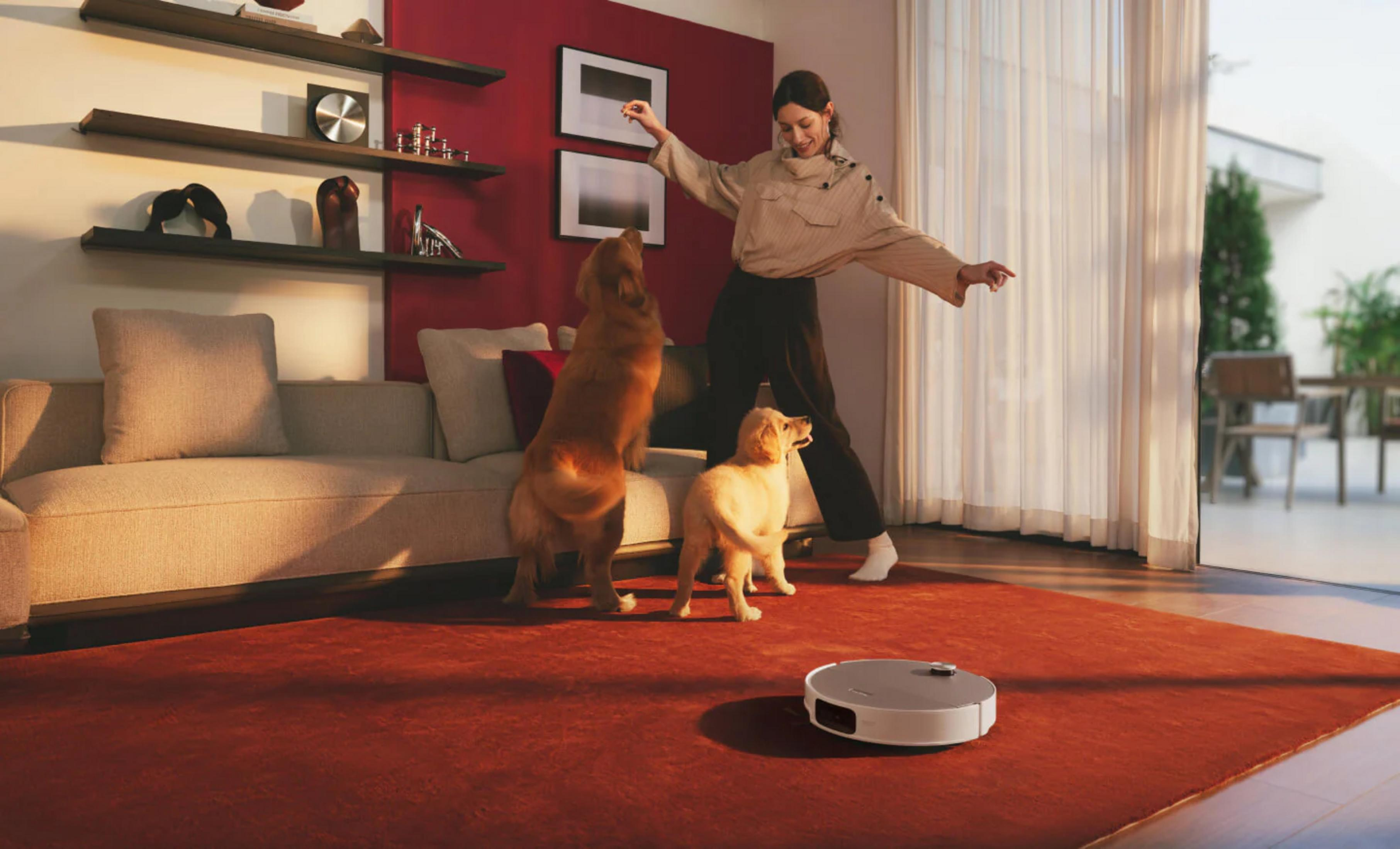 Una donna gioca con due golden retriever in un soggiorno con un robot aspirapolvere.