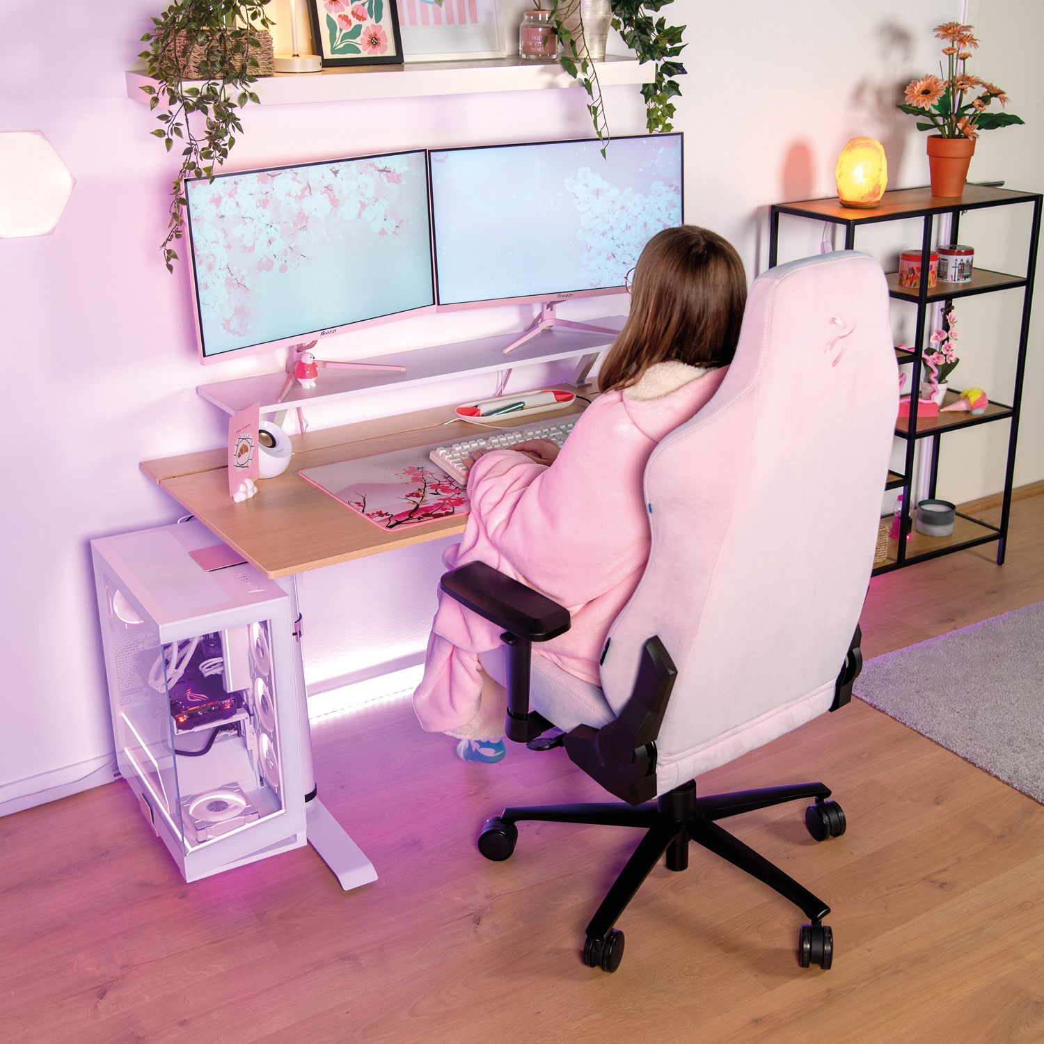 Una ragazza in tuta rosa è seduta a una scrivania con due monitor rosa, un PC bianco e una sedia rosa.