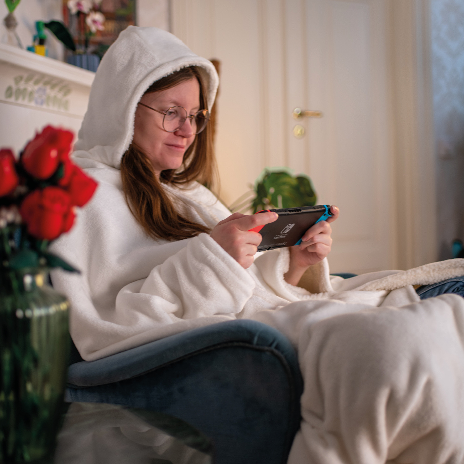 Donna in felpa bianca gioca a Nintendo Switch su poltrona blu, rose rosse in primo piano.