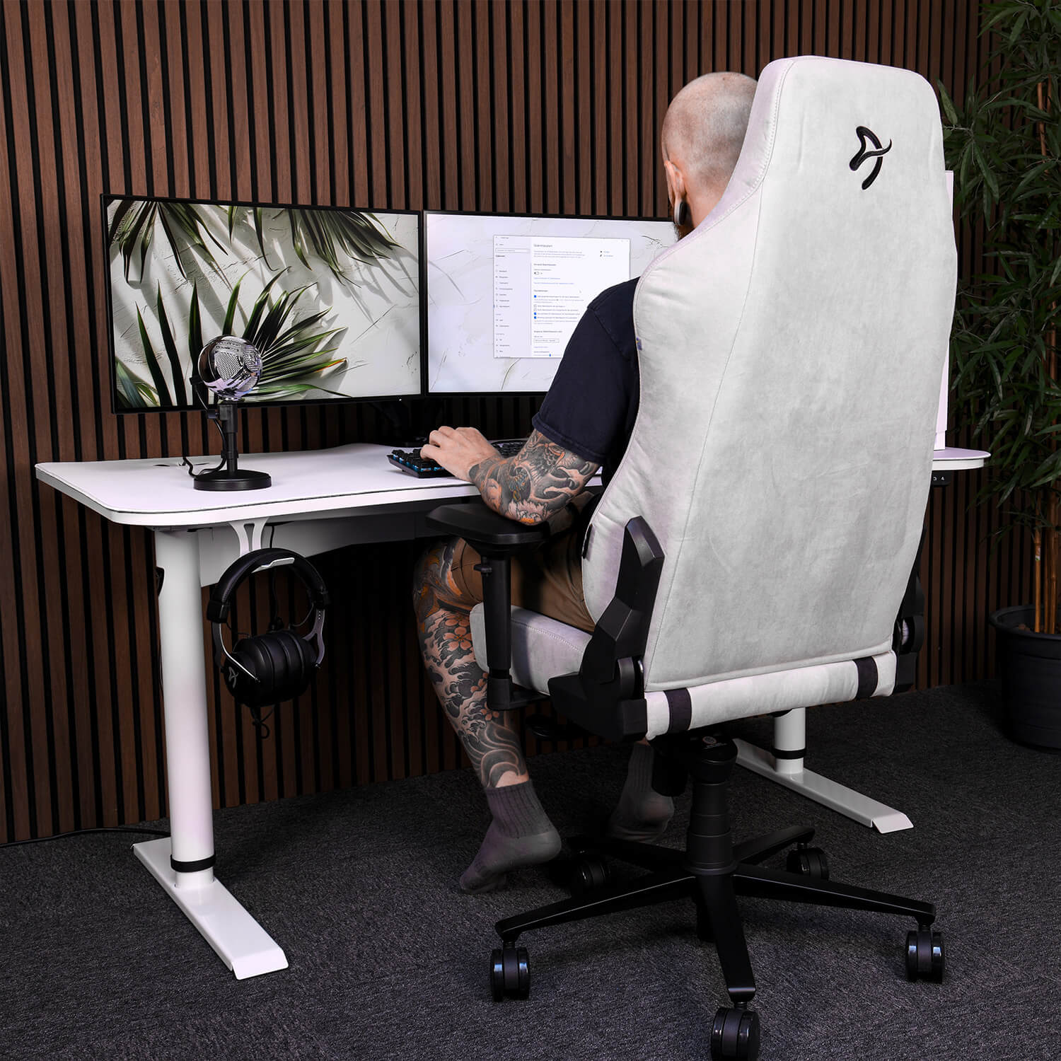 Uomo con tatuaggi seduto a una scrivania con due monitor e un microfono.