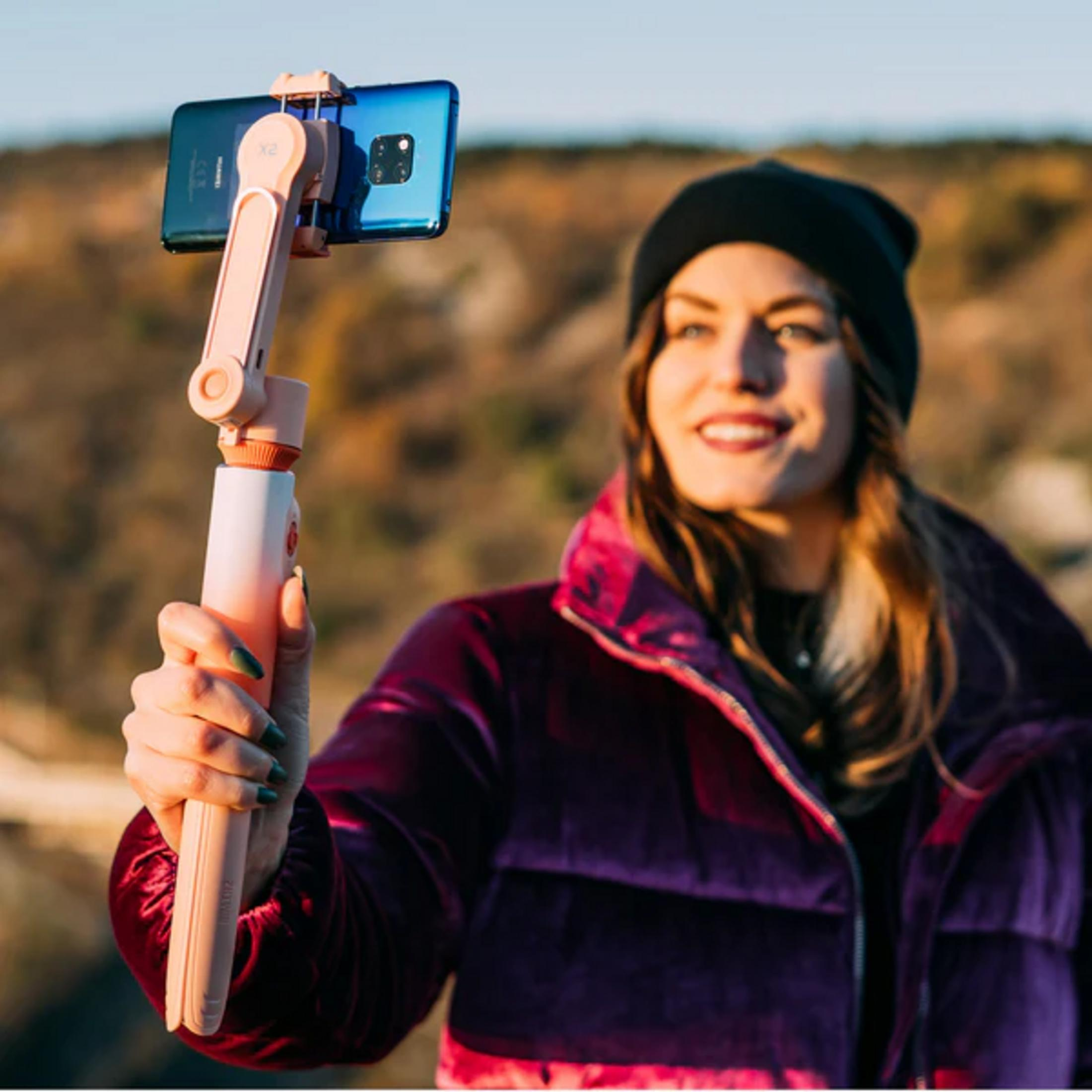 Eine Frau mit schwarzer Mütze und lila Jacke hält einen Selfie-Stick mit einem blauen Handy.