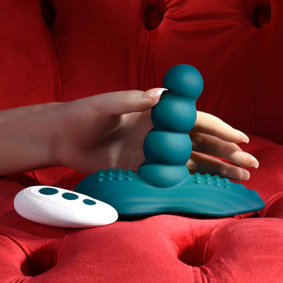 Eine Hand hält einen tealfarbenen Vibrator auf einem roten Samtstuhl. Ein weißer Controller liegt daneben.