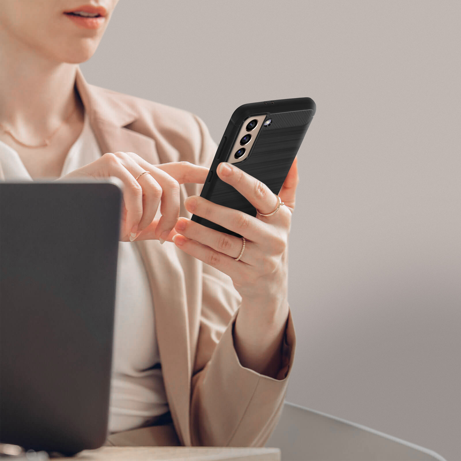 Donna in blazer usa uno smartphone con cover nera accanto a un laptop.