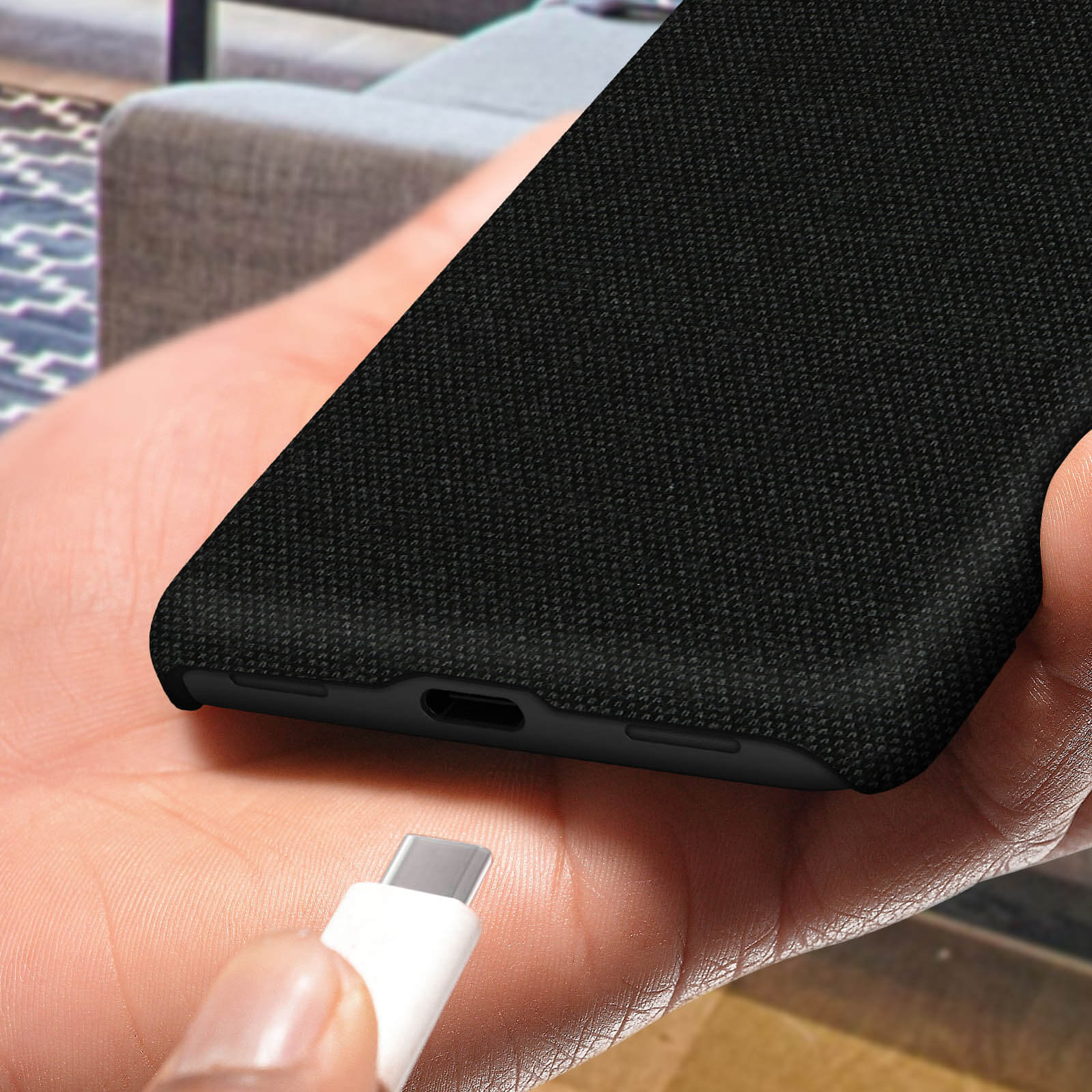 Une main tient une coque de téléphone noire avec un câble USB-C à proximité. Fond montre un canapé.