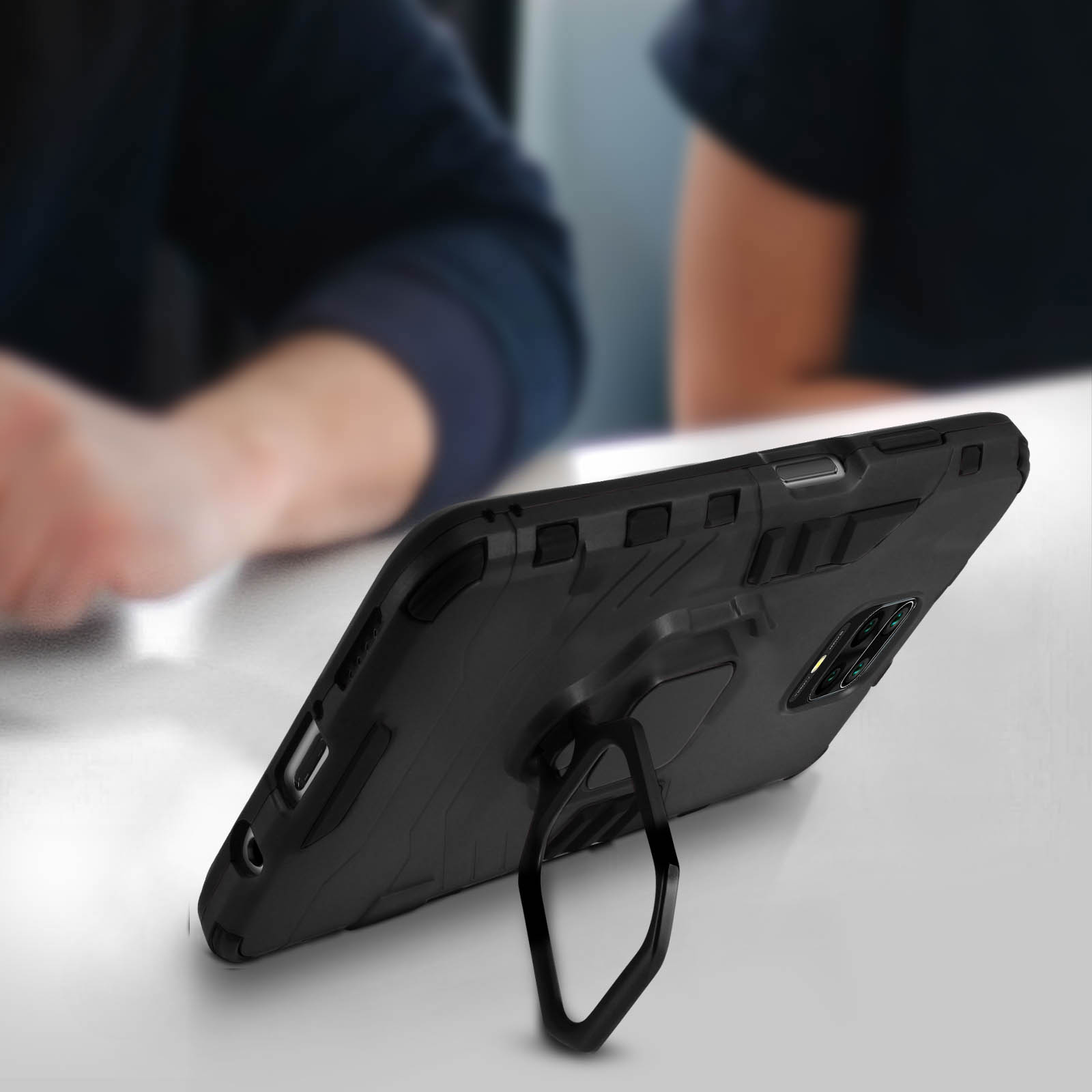 Custodia nera per telefono con anello di supporto su un tavolo, persone sullo sfondo.