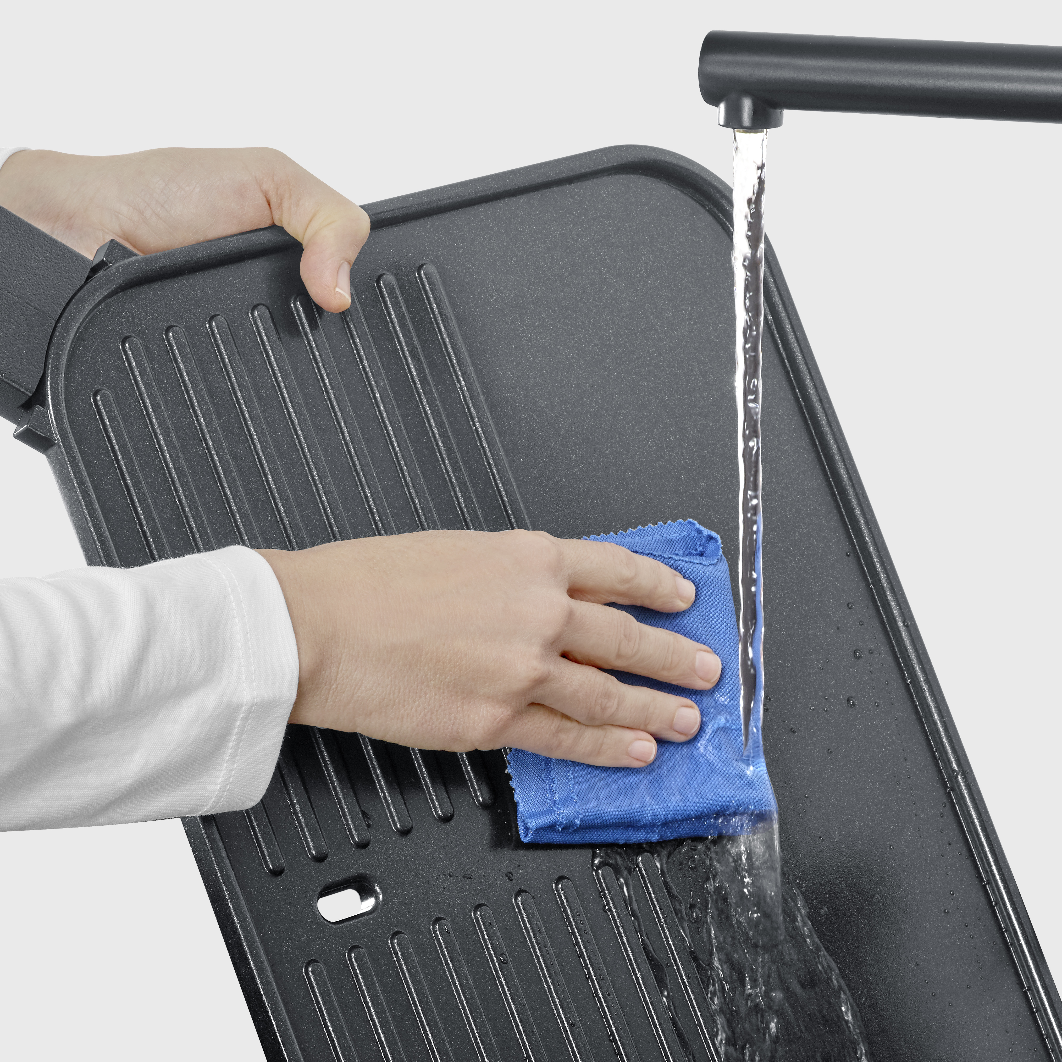 Una mano limpia una plancha con un paño azul bajo agua corriente.