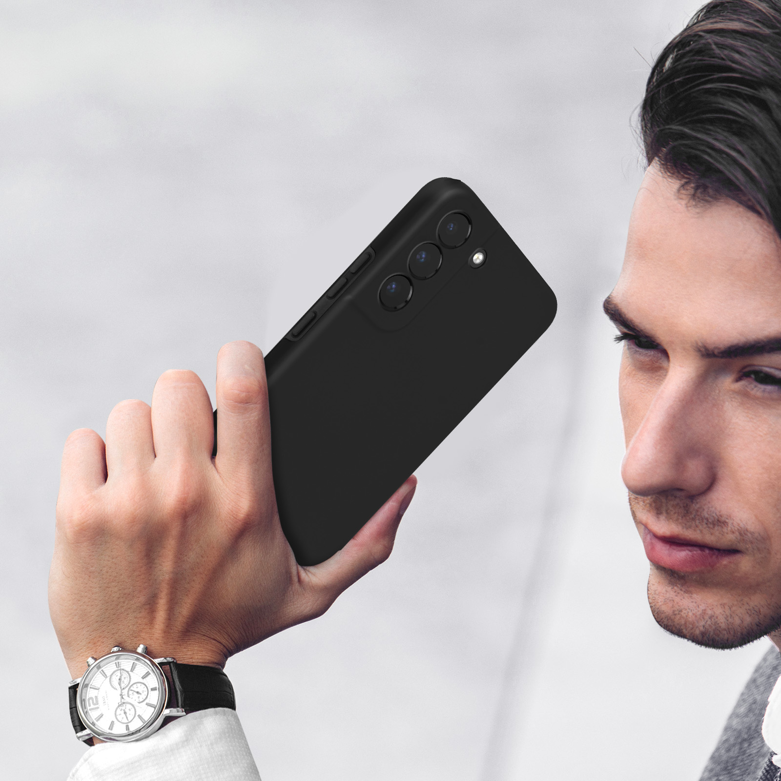 Homme tenant un téléphone noir à trois caméras. Montre à cadran blanc au poignet.