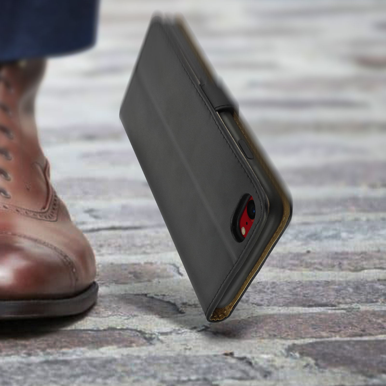 Una custodia nera per telefono poggia sui ciottoli accanto a una scarpa di pelle marrone.