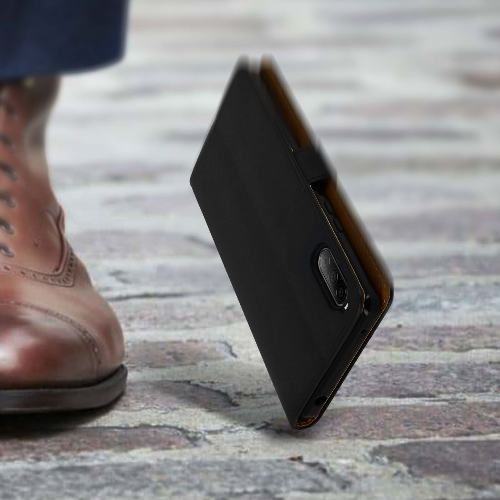 Una scarpa di pelle marrone calpesta vicino a un telefono con custodia nera su piastrelle.