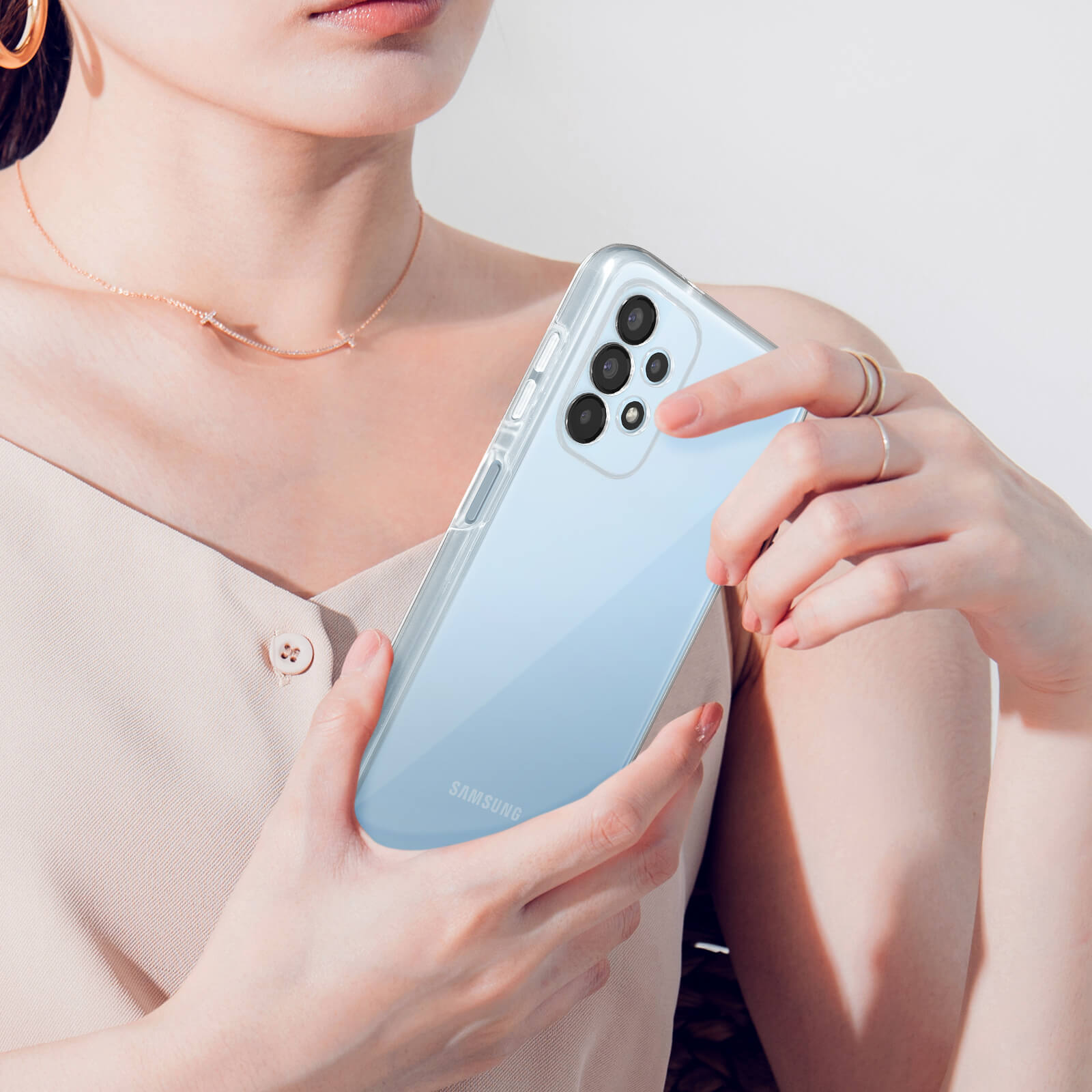 Una donna tiene un telefono Samsung azzurro con custodia trasparente. Indossa un top beige e una collana.