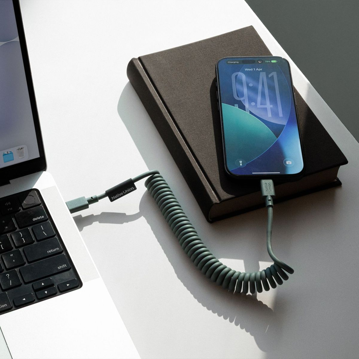 Un cavo grigio a spirale collega un telefono in carica su un libro a un laptop.