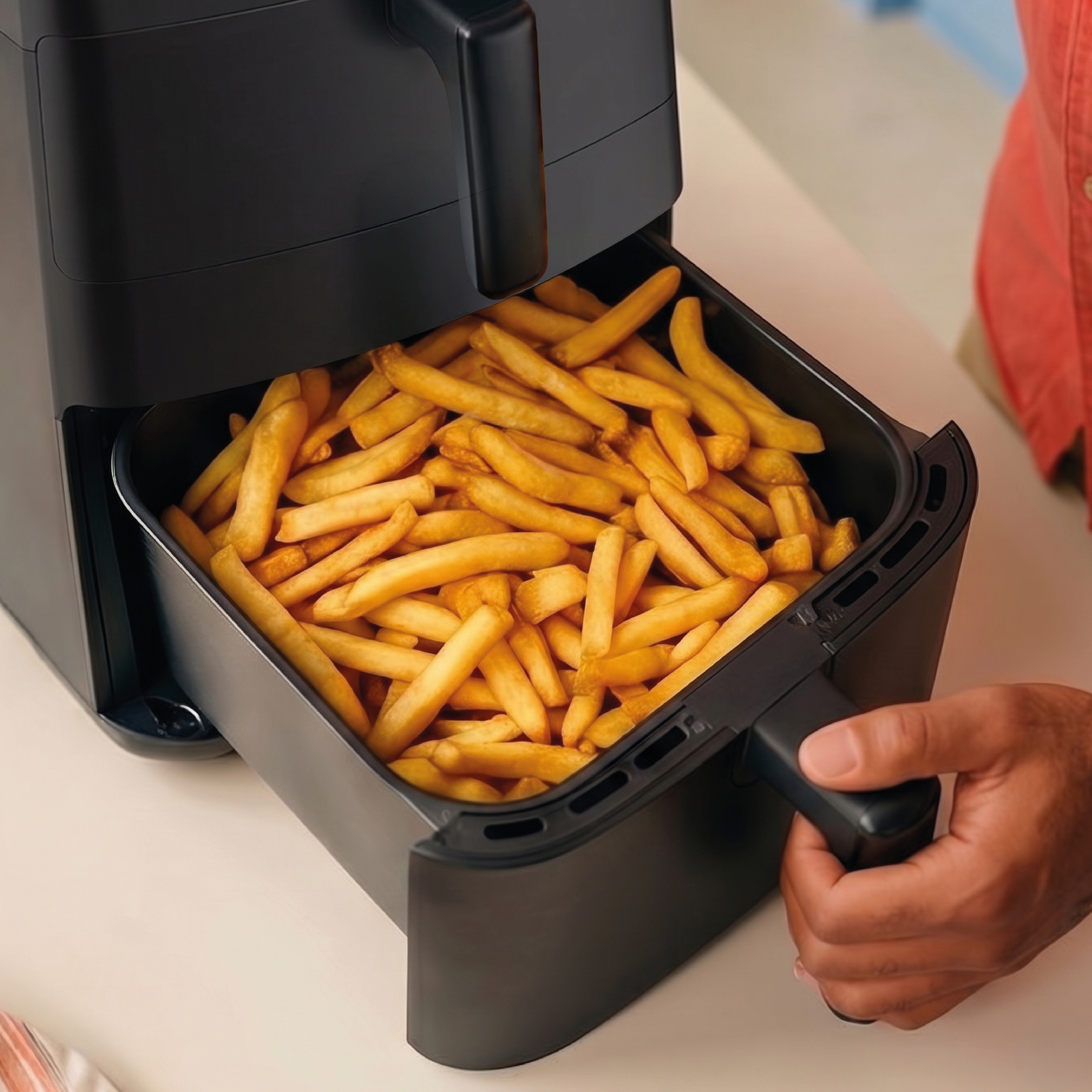 A black air fryer basket full of golden french fries is being pulled out.