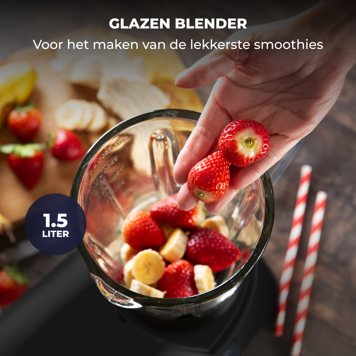 A hand adds strawberries and bananas to a glass blender. 1.5 liter capacity.