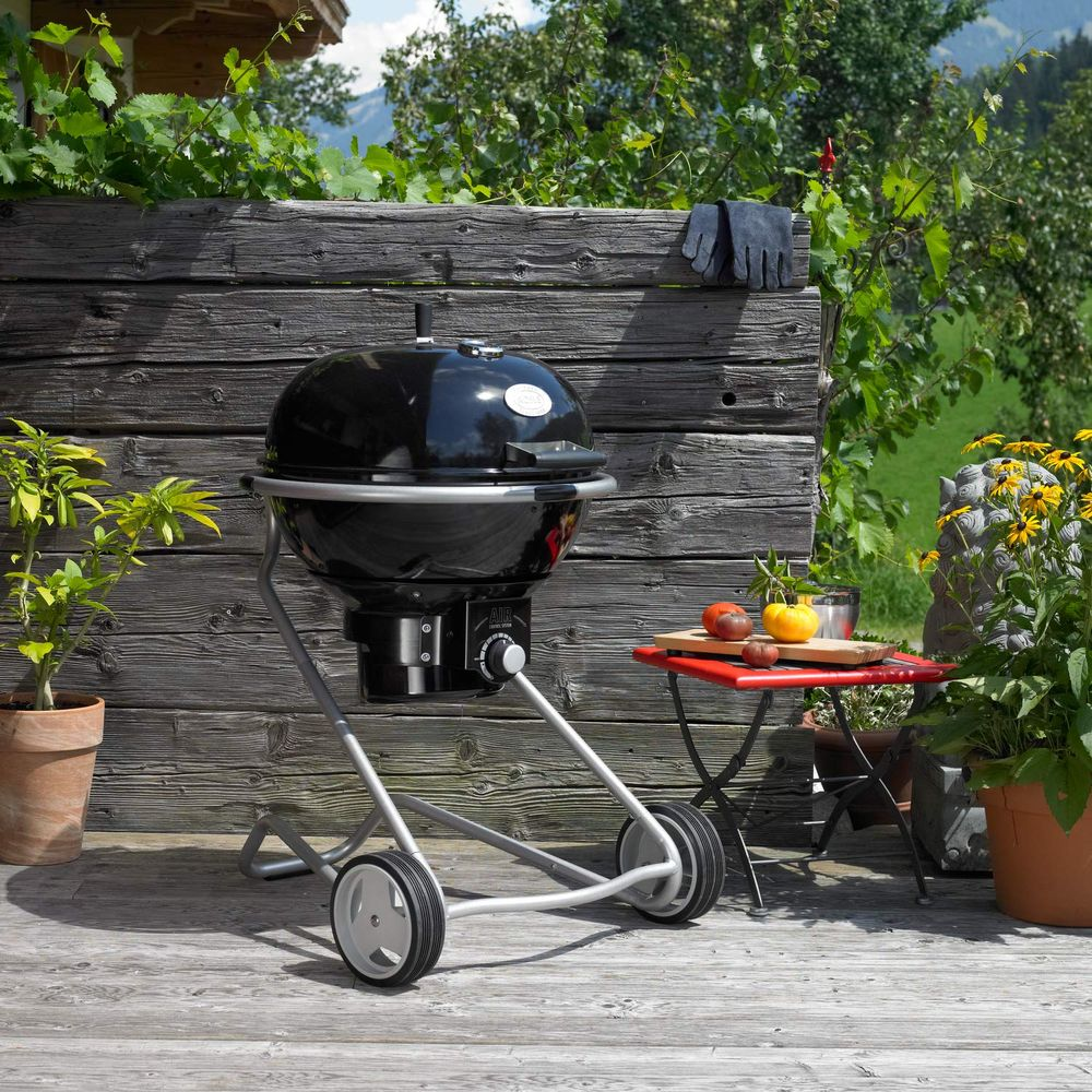 Ein schwarzer Grill mit silbernem Gestell steht auf einer Holzterrasse vor einer Holzwand.