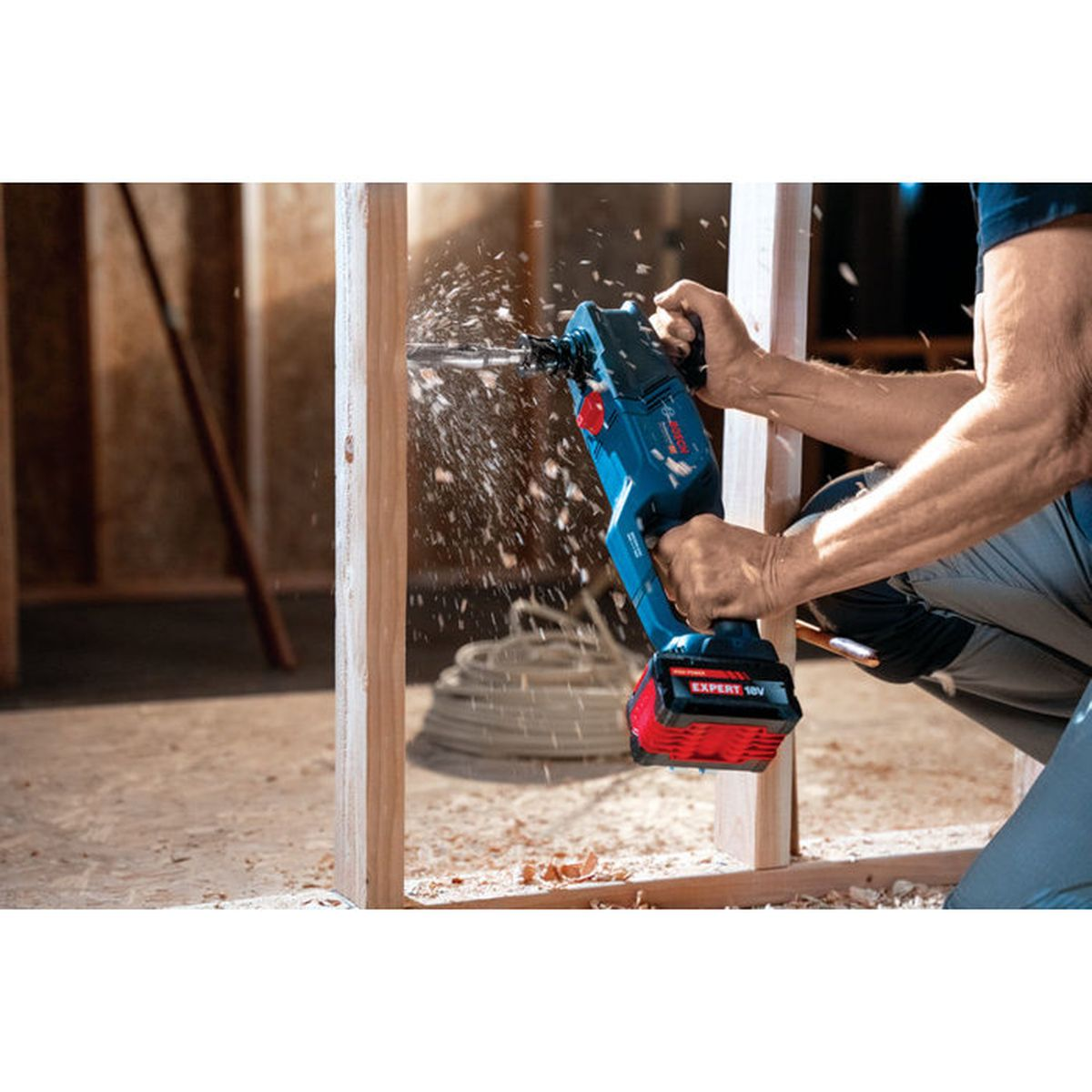 Una persona usa un taladro Bosch azul para taladrar un agujero en una viga de madera, creando serrín.