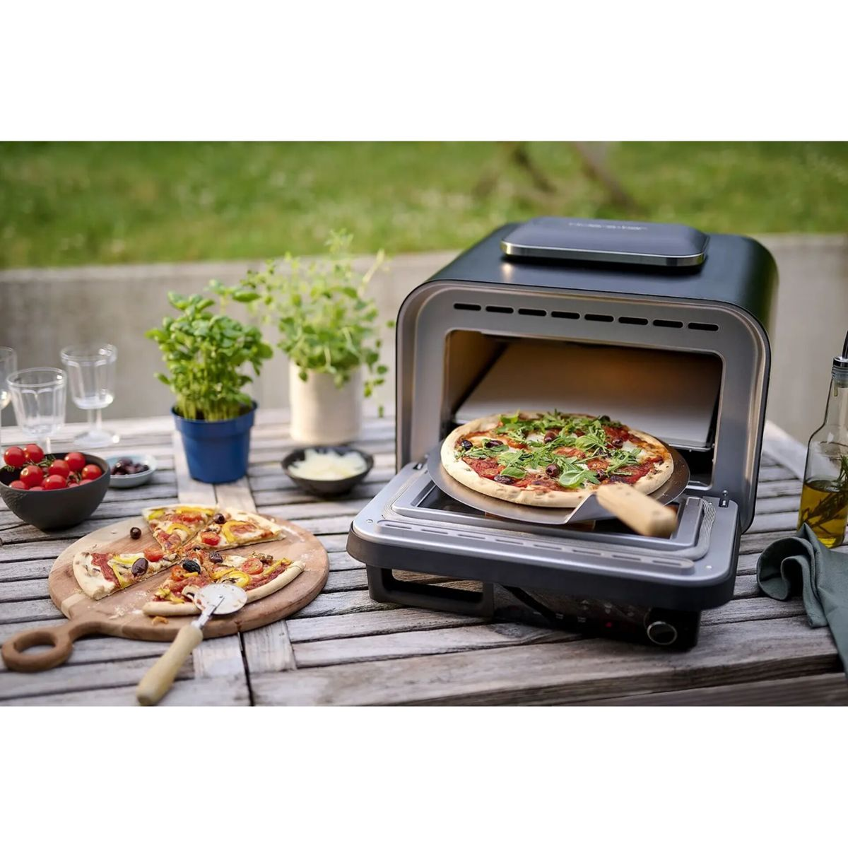 Pizzaofen im Freien mit einer Pizza, die herausgenommen wird, daneben eine angeschnittene Pizza.