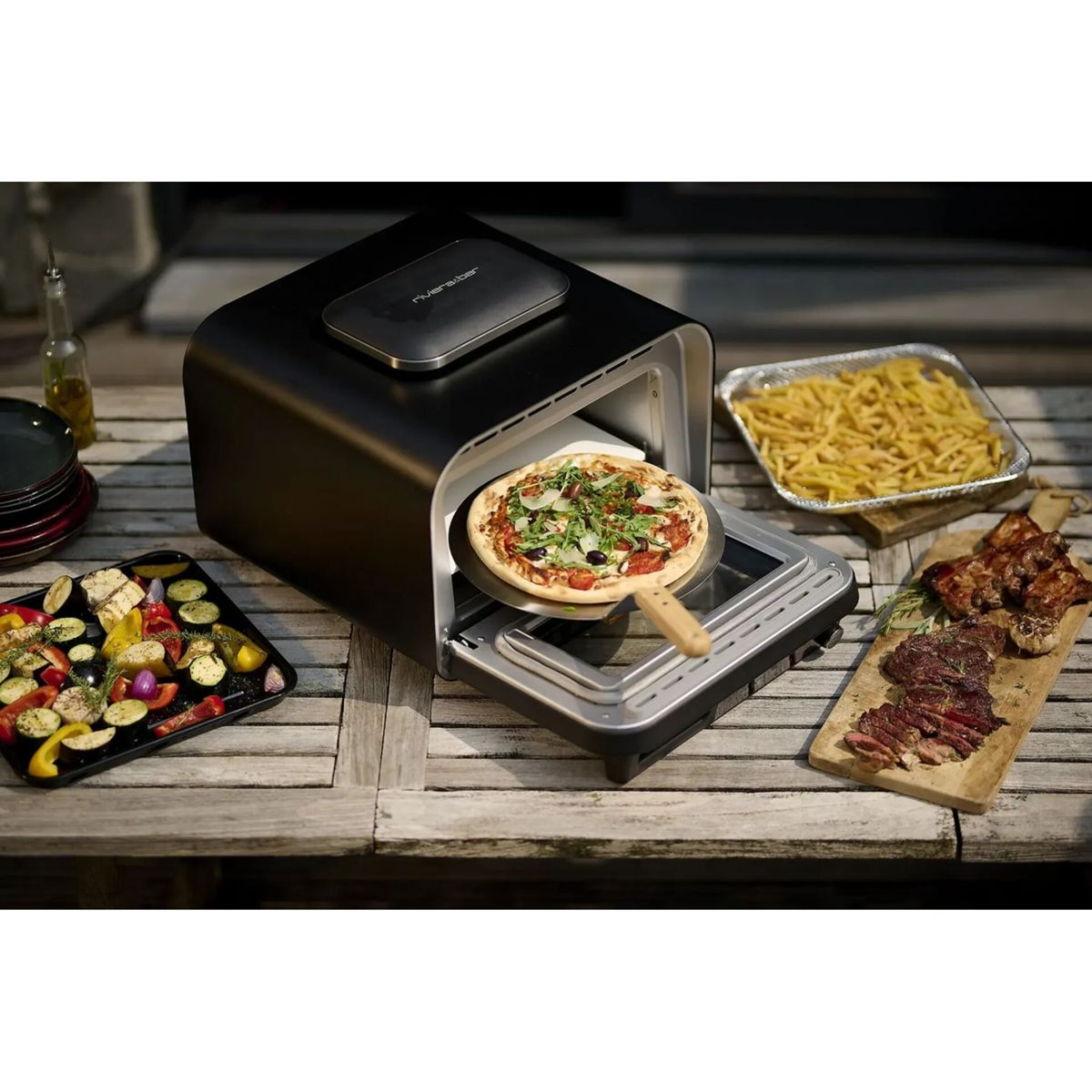 Ein schwarzer Pizzaofen mit einer Pizza auf dem Pizzaschieber, dazu Pommes, Gemüse und Fleisch.