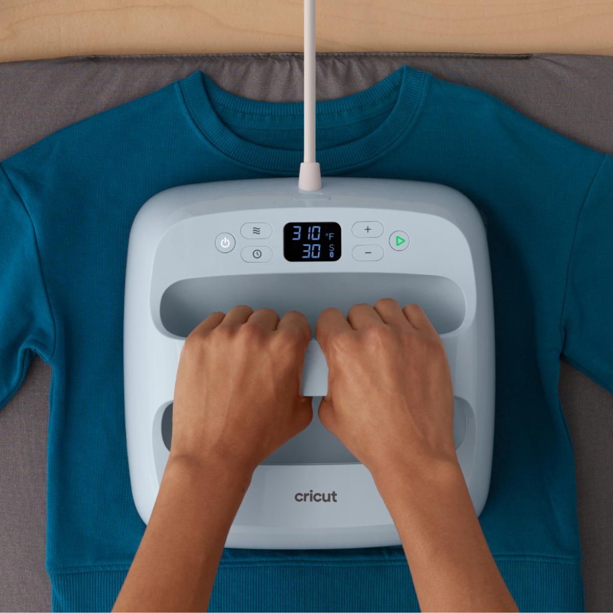 Mani premono una pressa a caldo Cricut azzurra su una felpa turchese.