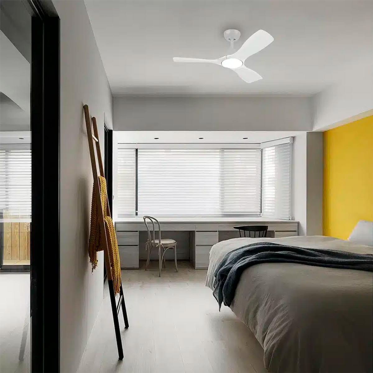 Camera da letto moderna con ventilatore a soffitto bianco, scrivania, letto con coperta blu, parete d'accento gialla.