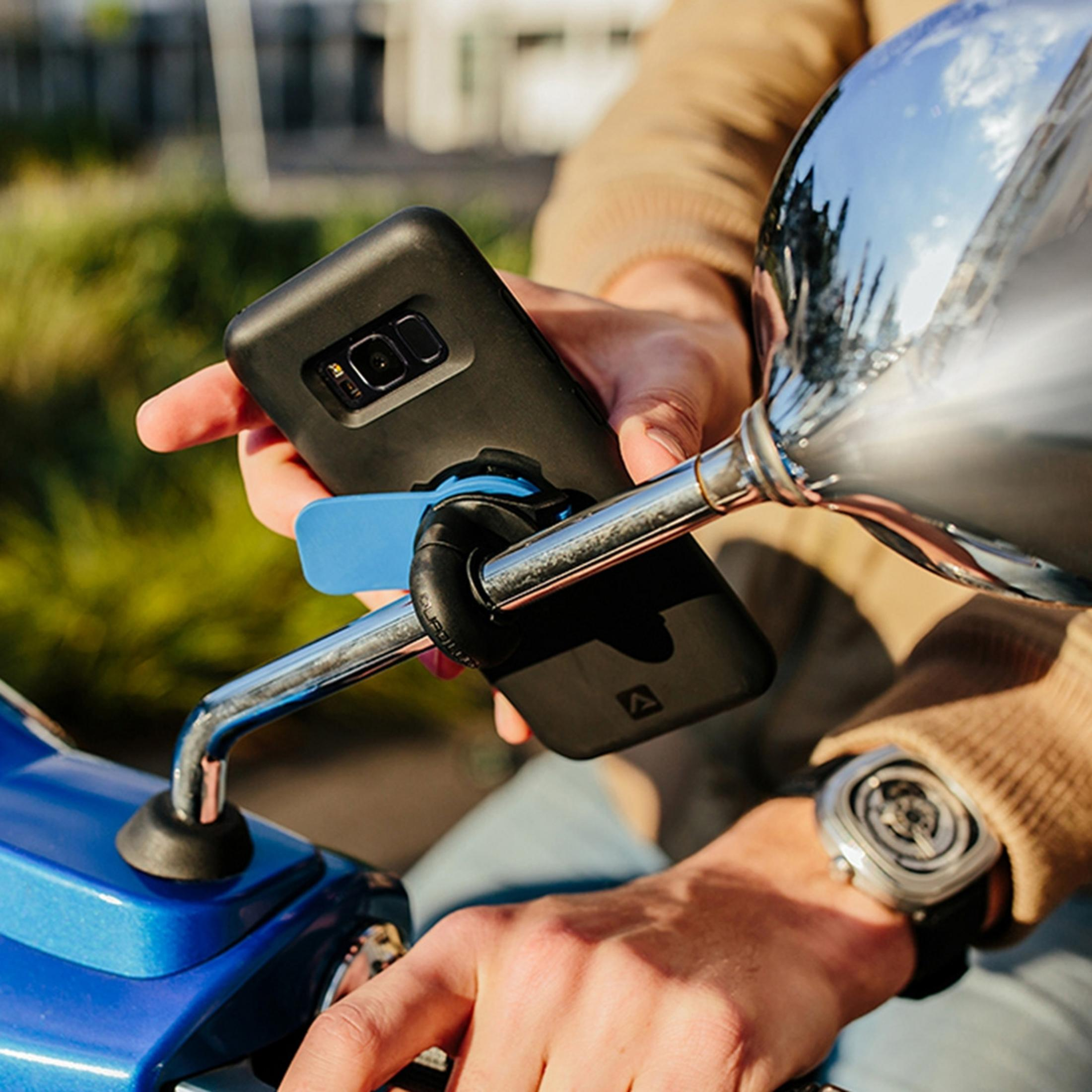 Person befestigt Handyhalterung am Motorrad-Spiegel. Blaue Halterung, schwarzes Handy.